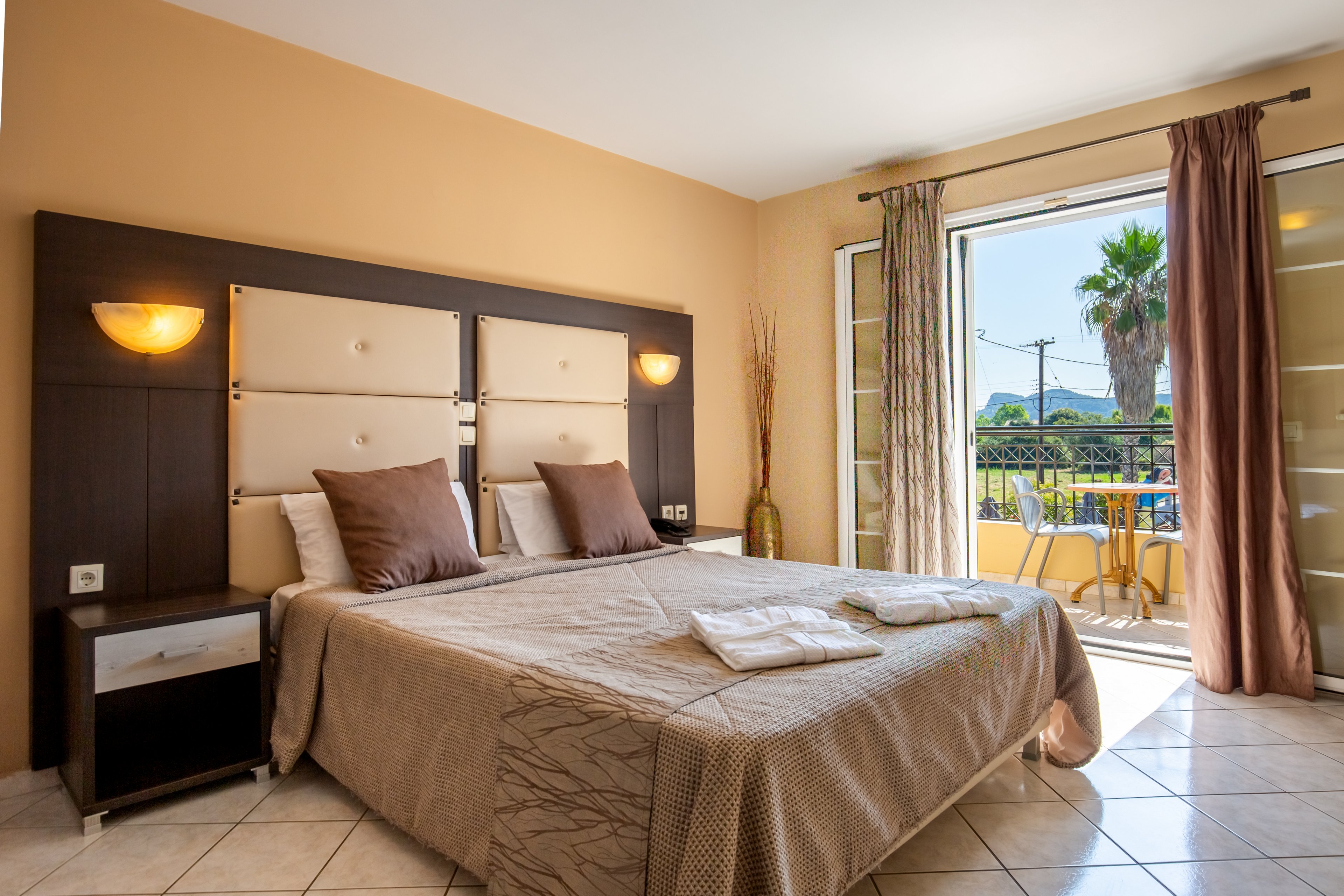 A neatly made double bed with neutral bedding and accent pillows sits in a bright hotel room. Warm wall sconces illuminate the padded headboard and bedside tables. The tiled floor leads to an open balcony door, revealing outdoor chairs, a table, and a palm tree in the distance. Sunlight streams through sheer curtains, creating an inviting and relaxing atmosphere.