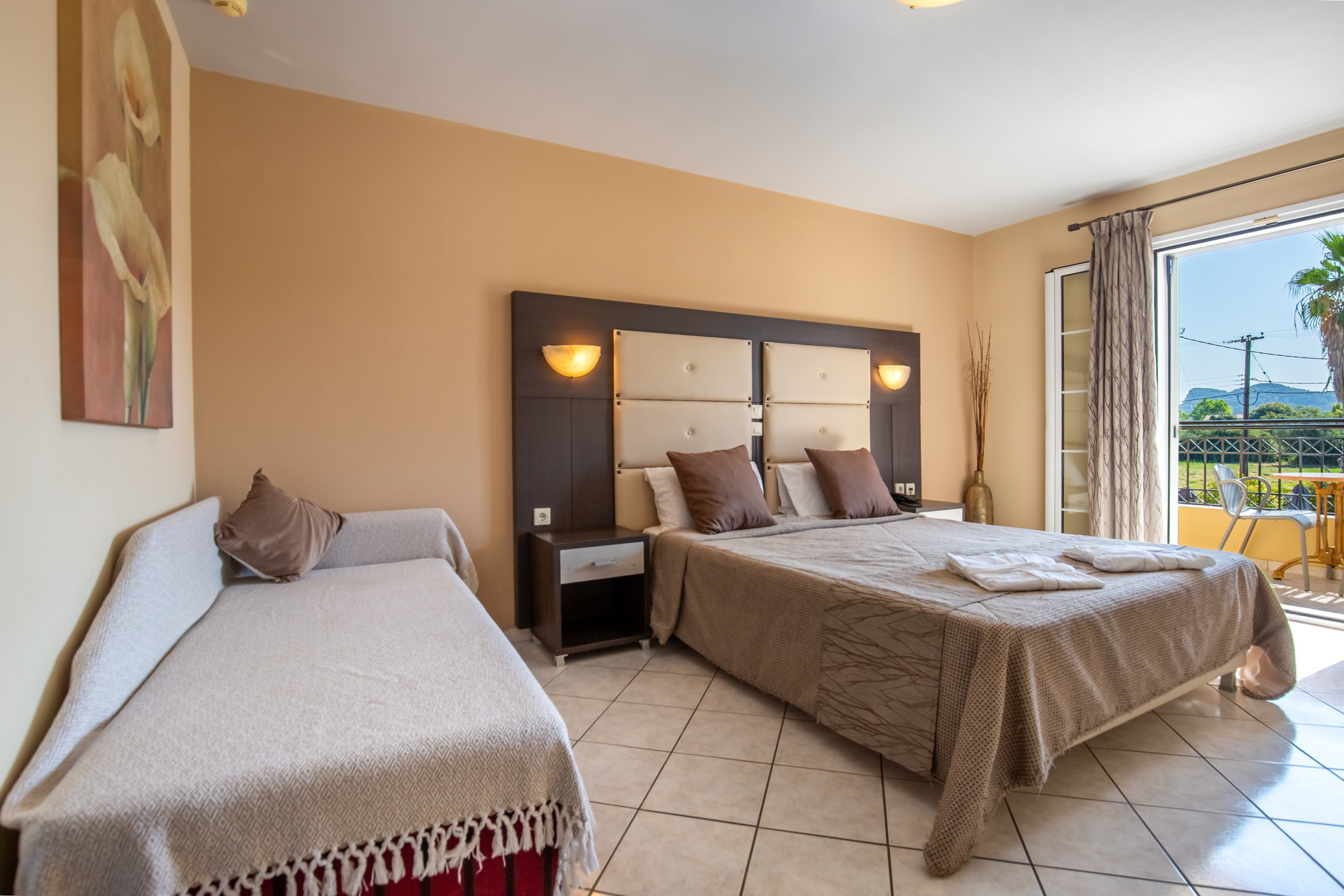 A spacious hotel bedroom features a large double bed with neutral bedding and multiple pillows. A smaller daybed sits to the side, topped with cushions and a fringed throw. Warm wall sconces flank a padded headboard, and a floral painting decorates the opposite wall. Sunlight enters through open glass doors leading to a balcony with outdoor furniture and a view of palm trees.