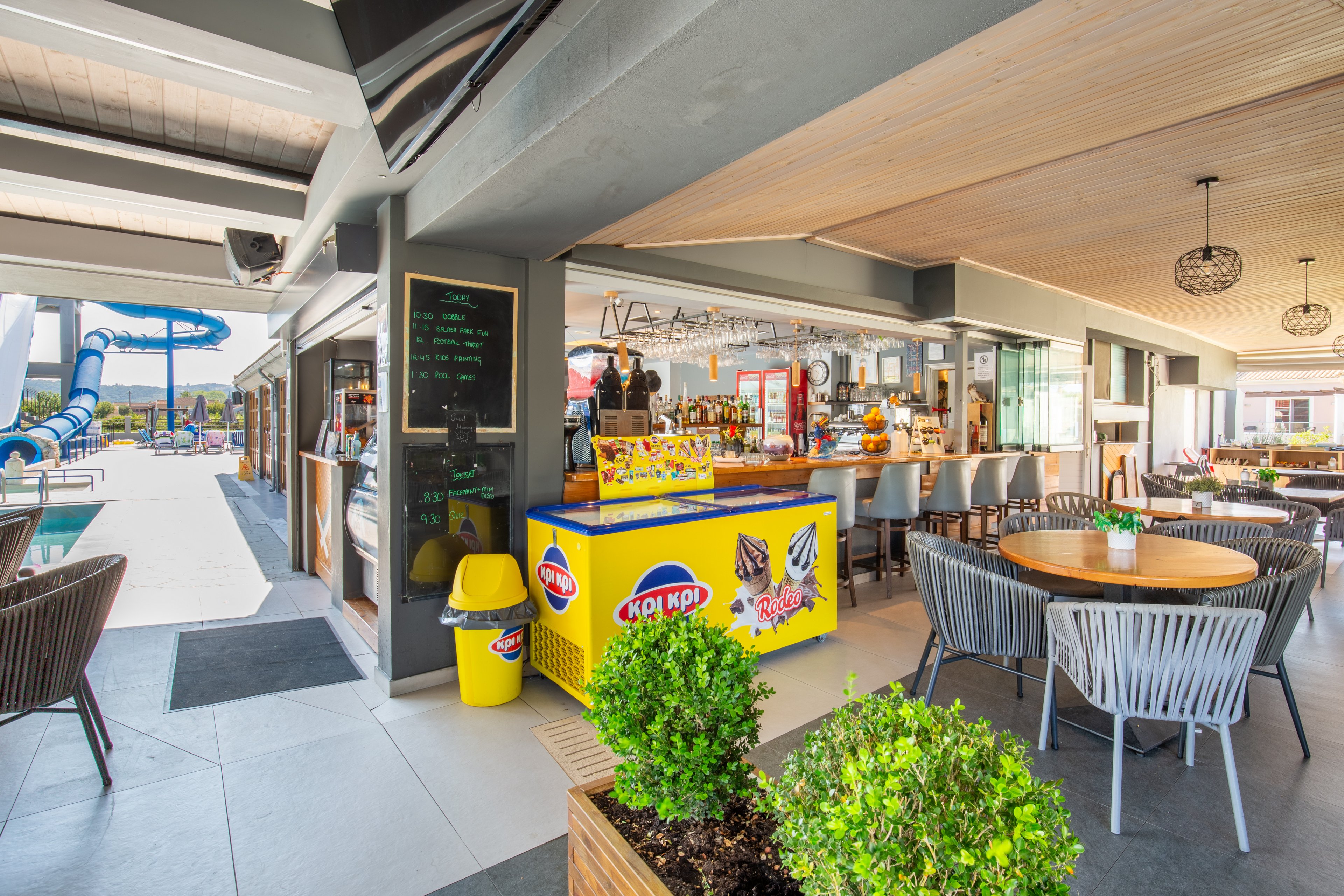A bright poolside snack bar is set in an open-air leisure area with modern tables and chairs. The central counter features a colorful ice cream freezer with visible product graphics and text. In the background, a waterslide and swimming pool indicate a family-friendly recreational setting. Potted green plants and warm wood accents create a relaxed, inviting atmosphere for guests.