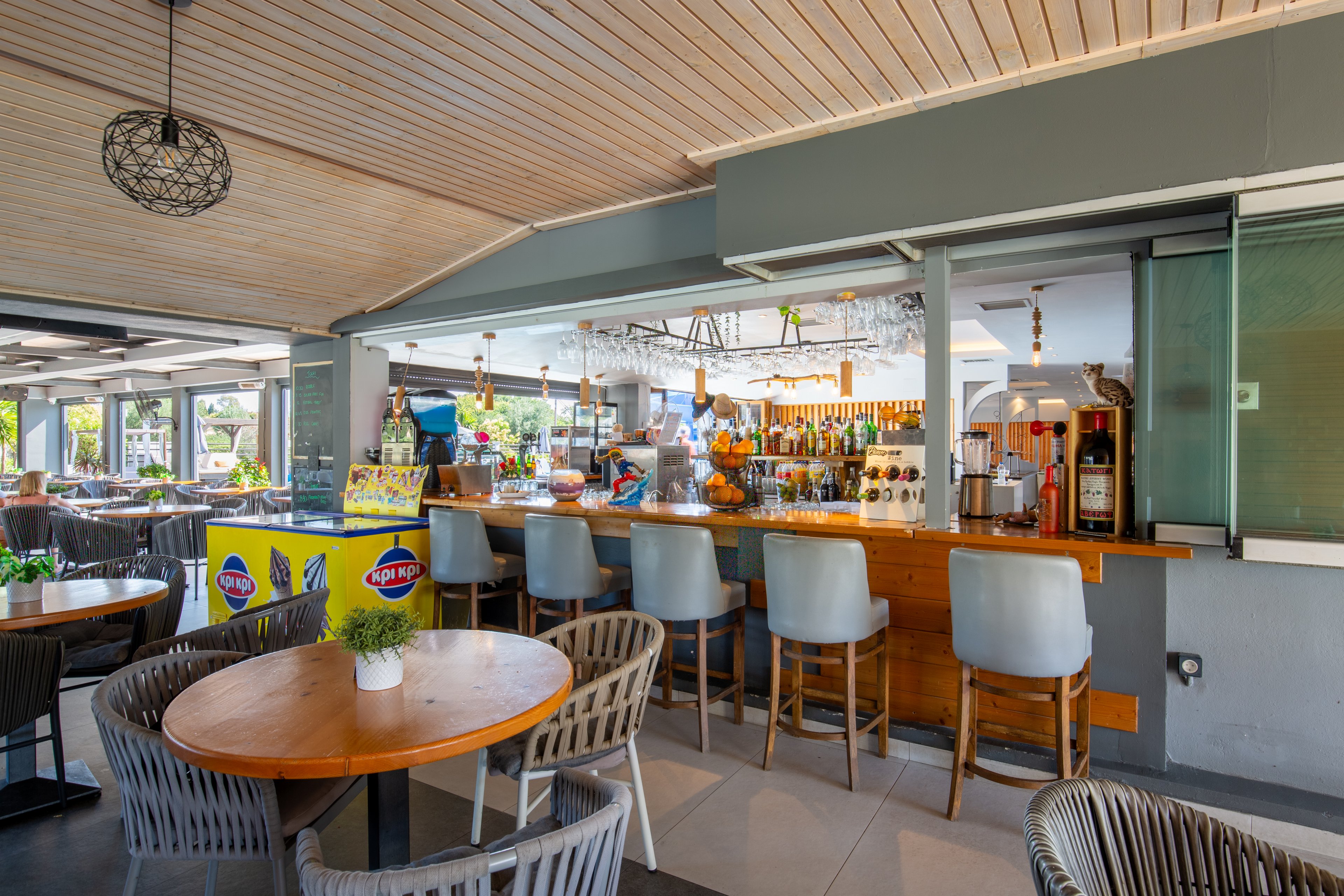 The image shows a modern open-air bar area with a wooden counter and high-backed stools. Round tables with woven chairs are arranged in the foreground, one featuring a small potted plant centerpiece. Behind the bar, shelves display various bottles and glassware under warm hanging lights. Large windows and an extended patio space create a relaxed, airy atmosphere suitable for casual dining and drinks.