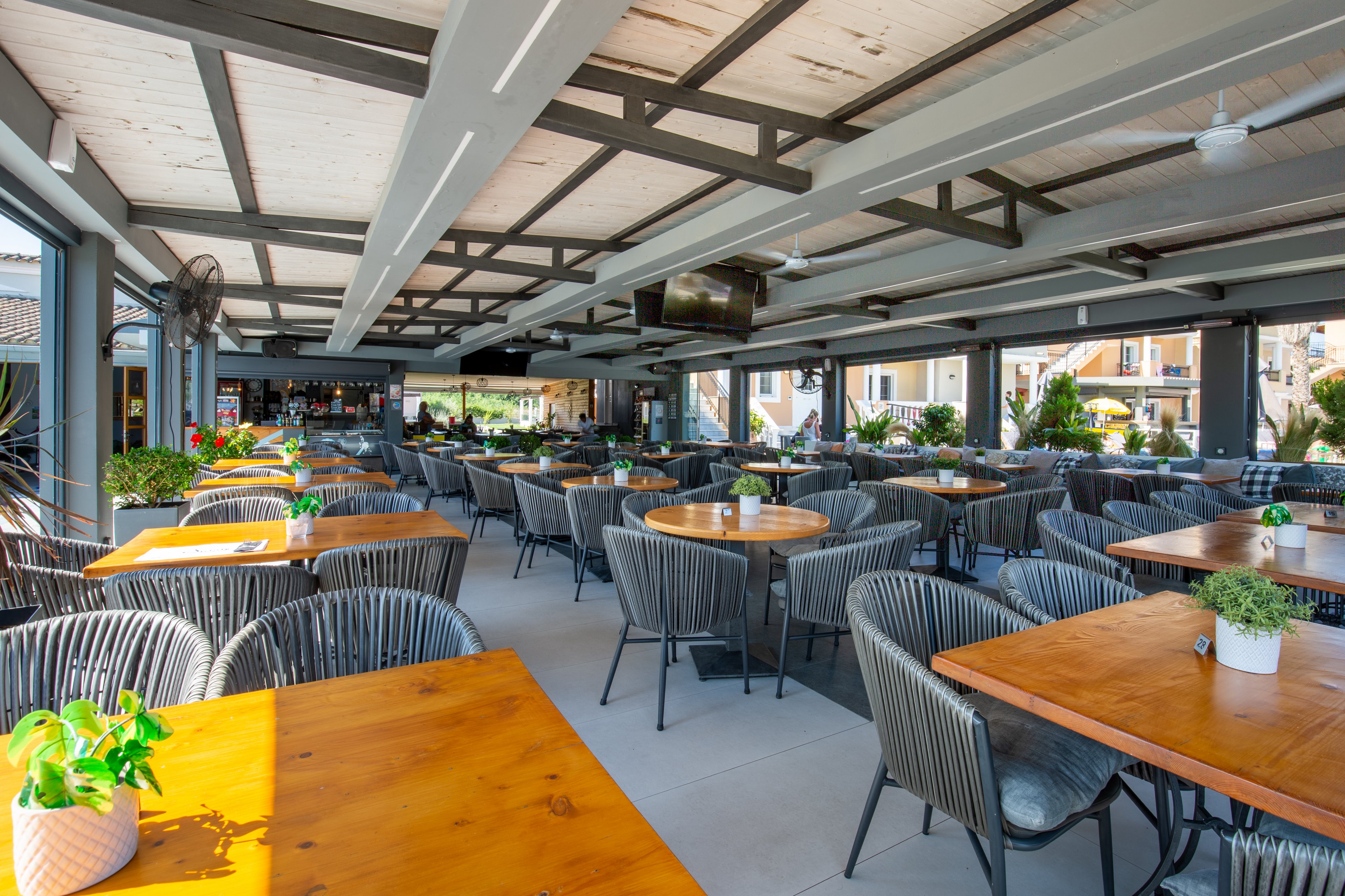 Spacious open-air dining area features rows of wooden tables and woven chairs arranged neatly under a covered terrace. The setting appears to be a modern restaurant or café with views of surrounding buildings and greenery. Potted plants decorate the tabletops, adding a touch of color and freshness to the scene. Bright daylight and clean architectural lines create an inviting, contemporary atmosphere ready for guests.