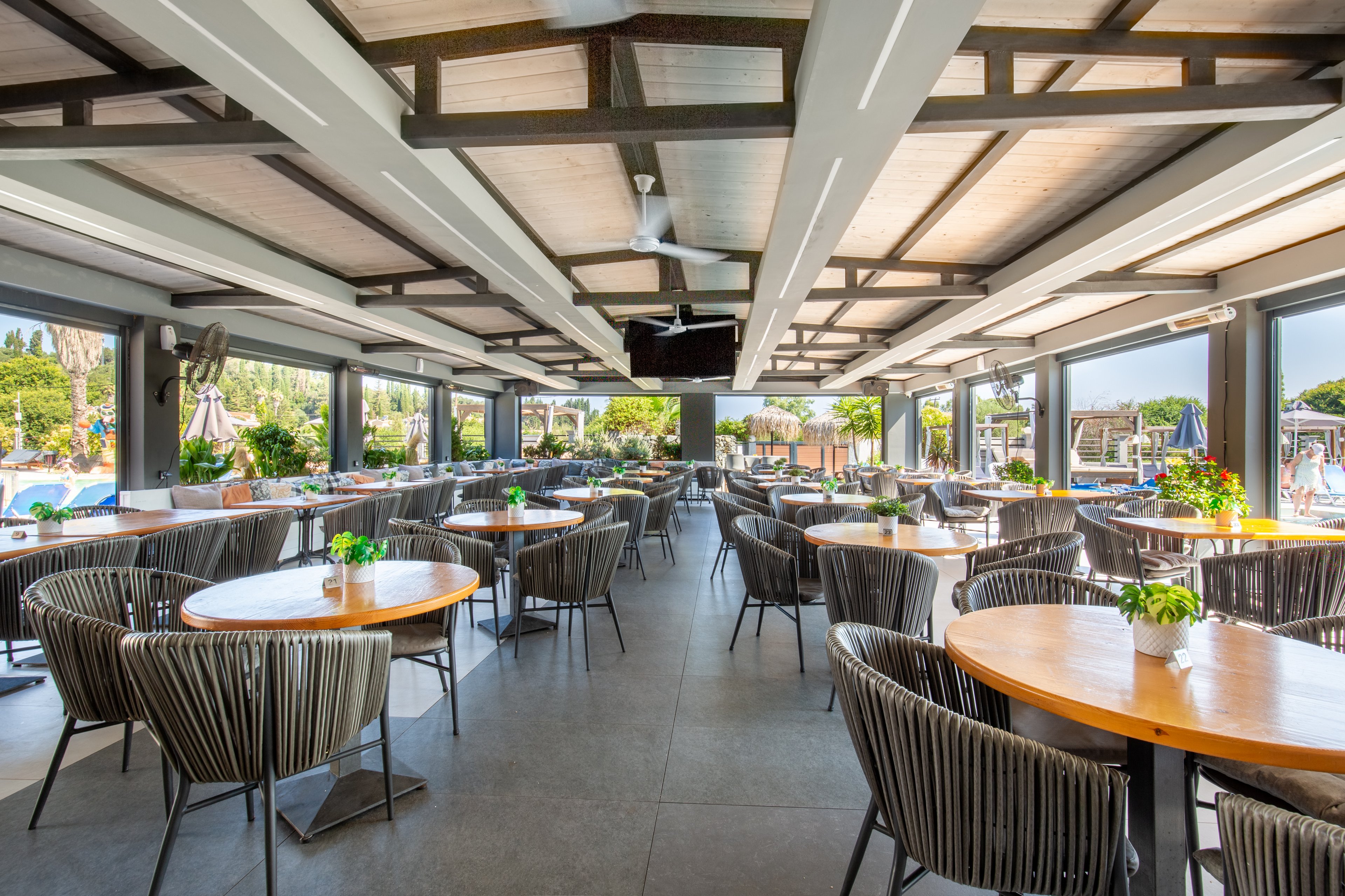 Spacious open-air restaurant dining area with numerous round wooden tables and modern woven chairs. The setting overlooks a resort pool and lush greenery, visible through large open sides. Potted plants decorate the tables, adding fresh color to the neutral interior. Exposed beams, ceiling fans, and natural light create a relaxed, contemporary vacation atmosphere.