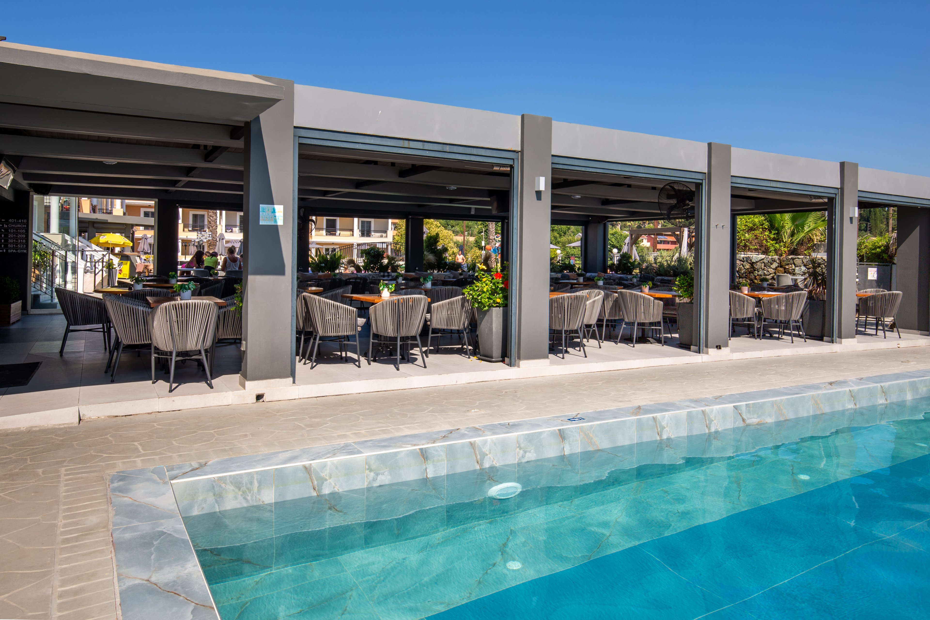 An open-air restaurant with shaded seating is situated beside a clear blue swimming pool. Modern gray columns and roof structures frame rows of tables and woven chairs. Green plants add color around the dining area, while nearby buildings are visible in the background. The bright daylight and blue sky create a relaxed vacation atmosphere.