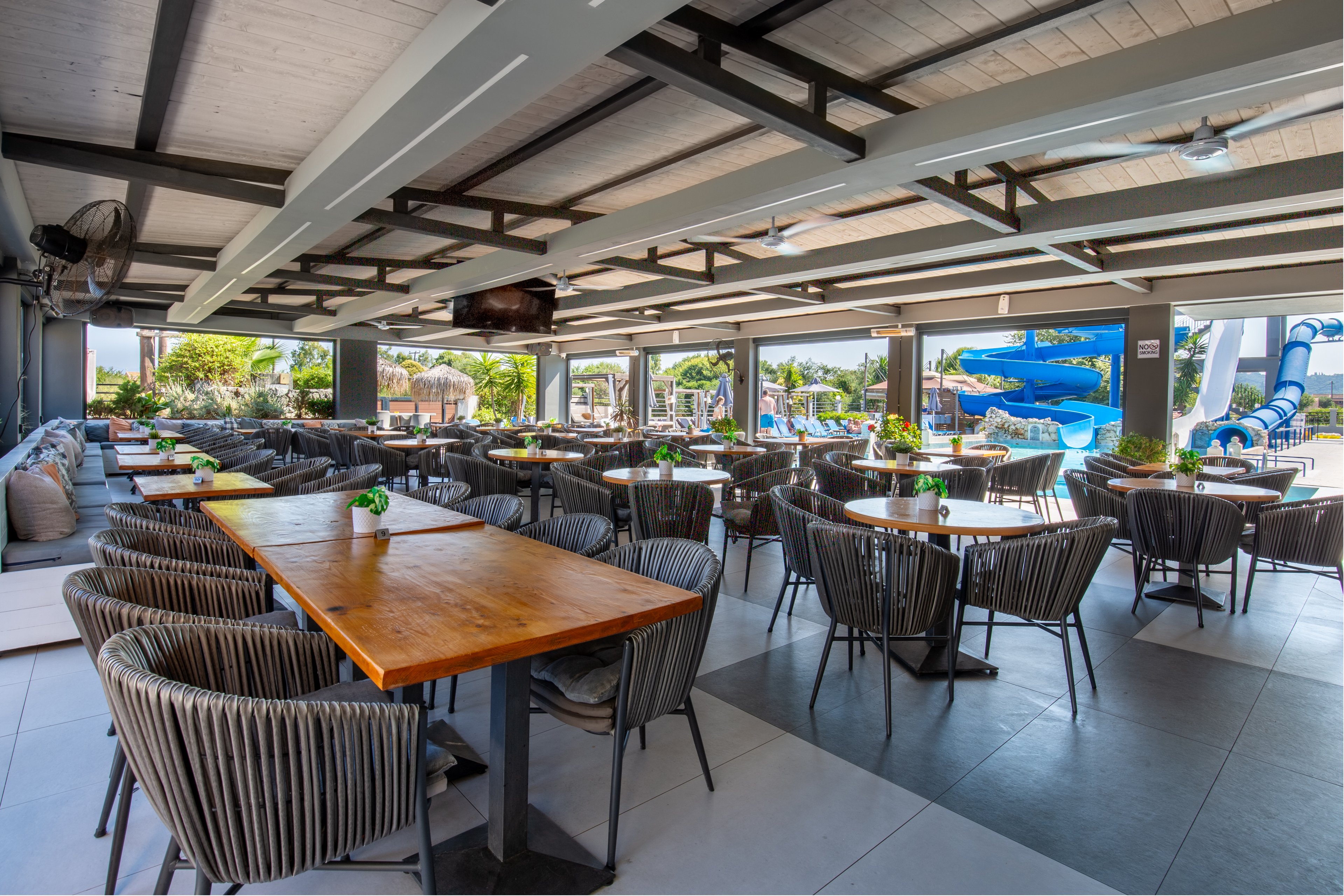Spacious open-air restaurant seating area with wooden tables and woven chairs is shown empty and ready for guests. Large windows and openings reveal an outdoor water park with slides and pools in the background. Neutral gray flooring and ceiling beams contrast with warm wood tabletops and green plant centerpieces. The scene suggests a relaxed vacation atmosphere at a coastal or resort property.