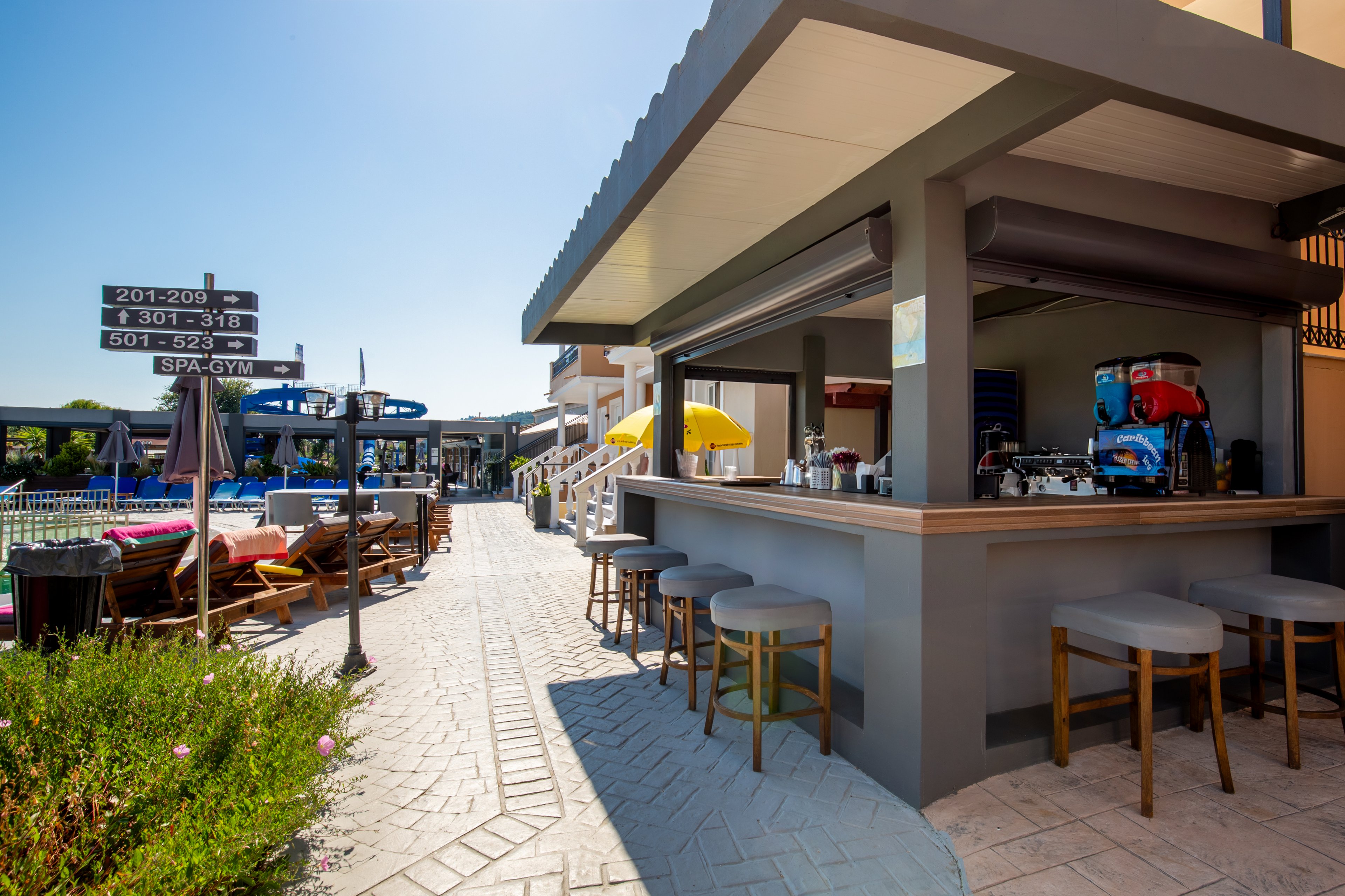 An open-air bar with stools sits along a paved walkway in a resort setting. Sun loungers and umbrellas line the pool area in the background under a clear blue sky. A directional signpost indicates room numbers and a spa and gym further down the path. Bright colors from umbrellas and outdoor furniture create a relaxed vacation atmosphere. The scene suggests a leisure destination designed for comfort and casual socializing.