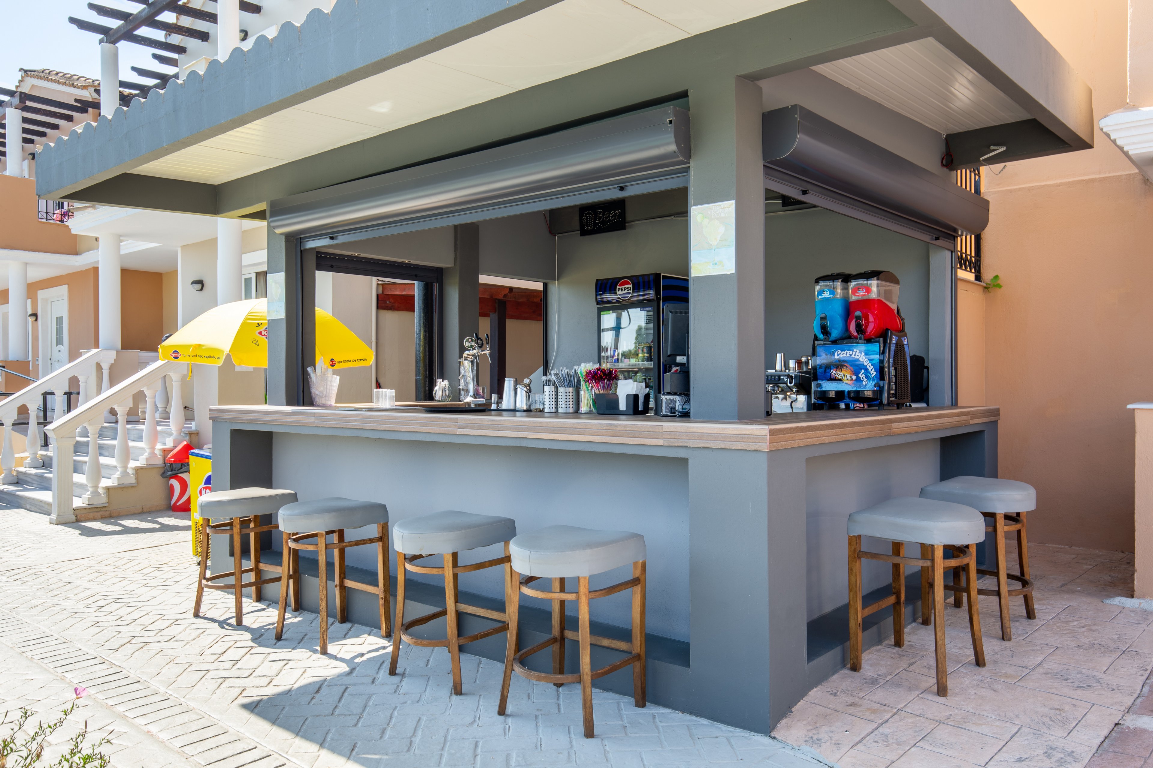 An open-air bar counter stands in a sunny resort courtyard, surrounded by cushioned wooden stools. The bar area features drink dispensers, glassware, and a refrigerator inside the structure. Nearby, a bright yellow umbrella and white staircases lead up to adjacent buildings, enhancing the vacation atmosphere. The scene is clean, inviting, and designed for relaxed outdoor refreshments.
