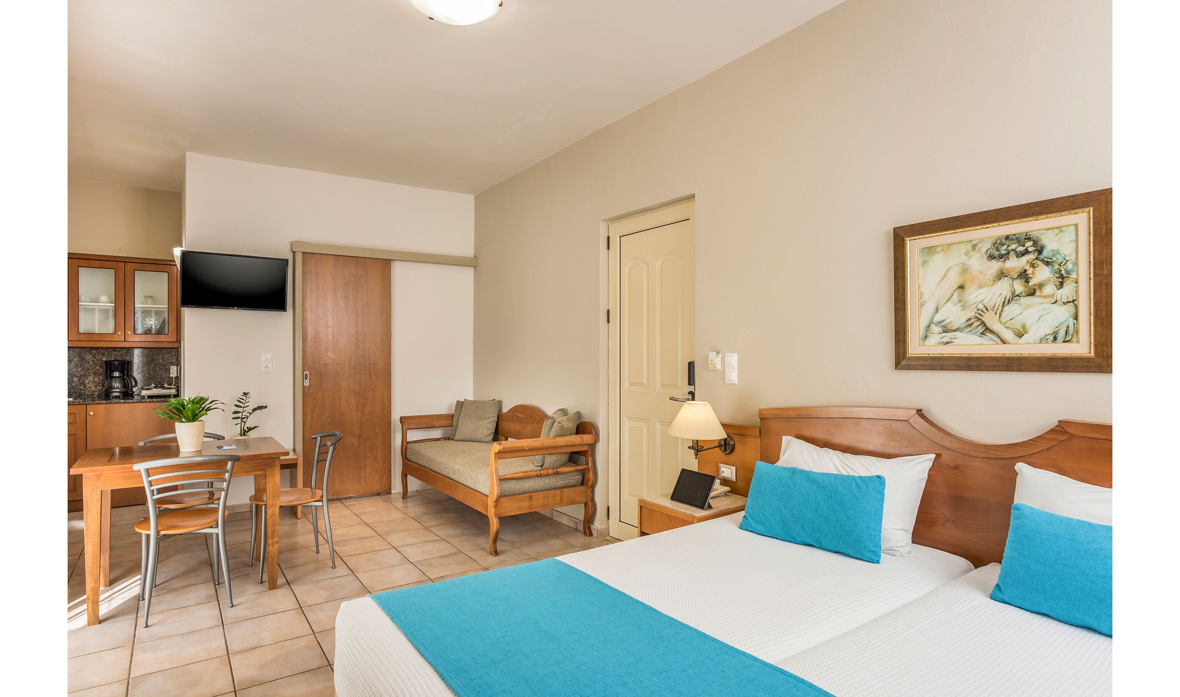 A bright, tidy hotel room features two twin beds with white linens and turquoise bed runners. The space includes a small wooden dining table with metal chairs and a wall-mounted television near a kitchenette area. A wooden sofa with beige cushions sits against the wall beside a door, and a bedside lamp adds warm light. Neutral walls, tiled flooring, and a framed artwork create a calm, welcoming atmosphere.