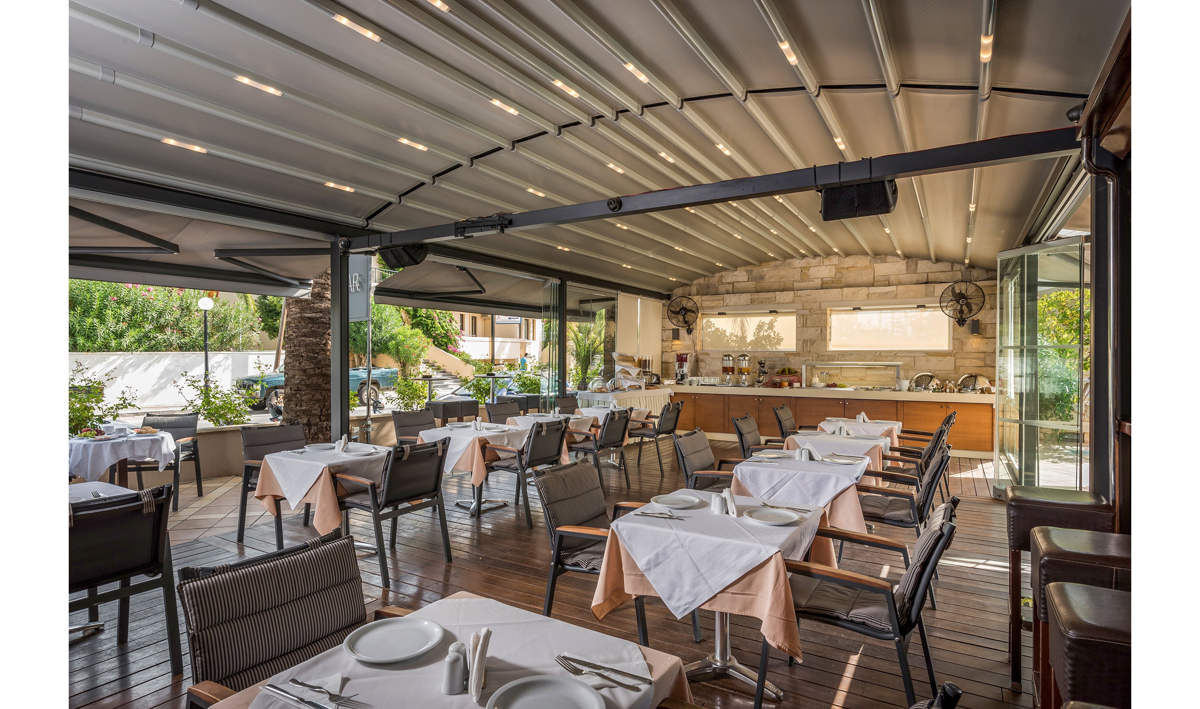 The image shows an open-air restaurant dining area with neatly arranged tables and chairs. Each table is set with white tablecloths, plates, glasses, and cutlery, ready for guests. A long counter and buffet area are visible at the back, framed by stone walls and large windows. The space features a covered terrace roof and overlooks greenery, creating a relaxed and inviting atmosphere.