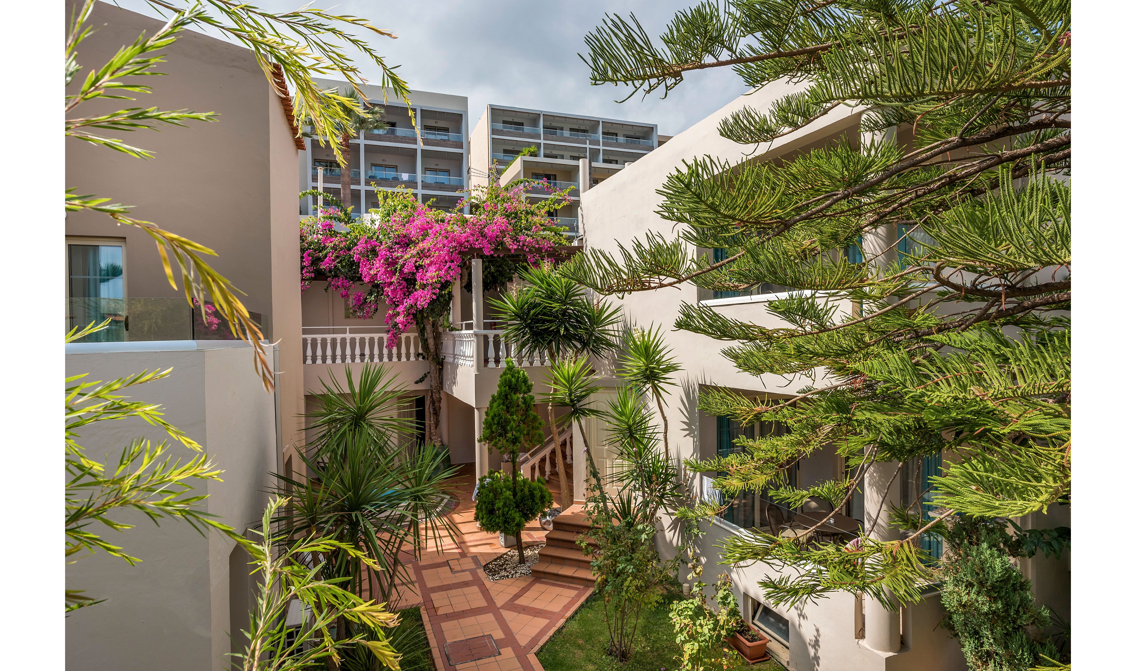 A sunlit courtyard is framed by modern beige buildings and vibrant greenery. Tall tropical trees and potted plants line the tiled walkway, leading toward balconies and staircases. Bright pink bougainvillea cascades over a terrace, adding color against the neutral architecture. The scene suggests a tranquil hotel or apartment complex in a warm coastal climate.