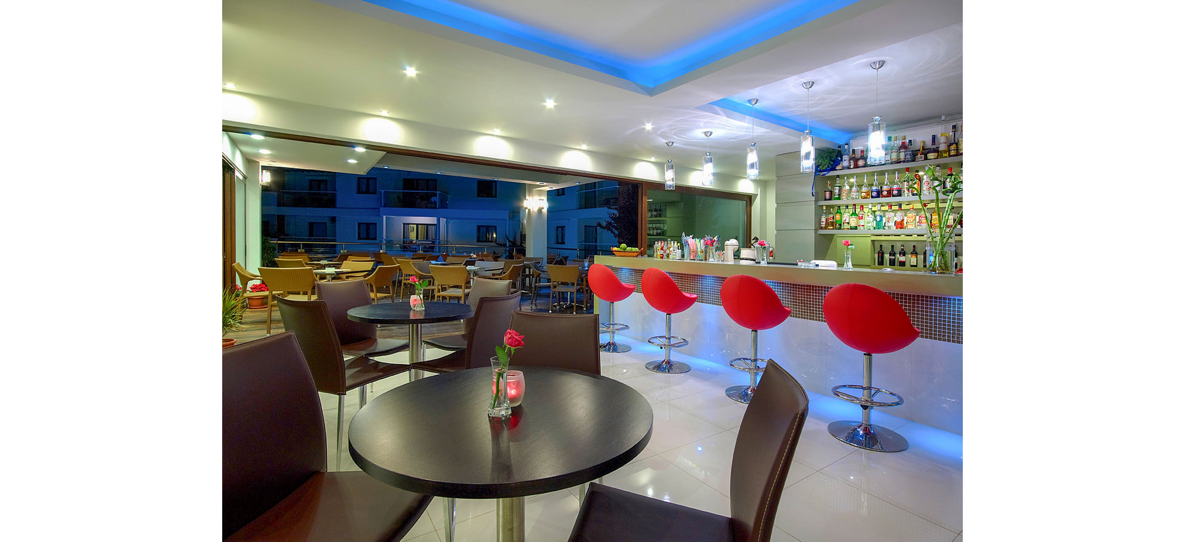 Contemporary bar lounge interior featuring round tables and red bar stools. The setting includes a well-stocked back bar with bottles and glassware, illuminated by pendant lights and blue ceiling accent lighting. Large windows open to an outdoor seating area with additional tables and chairs, suggesting a relaxed social atmosphere. The polished floor, sleek furniture, and small flower vases on tables create a stylish, inviting environment for guests.