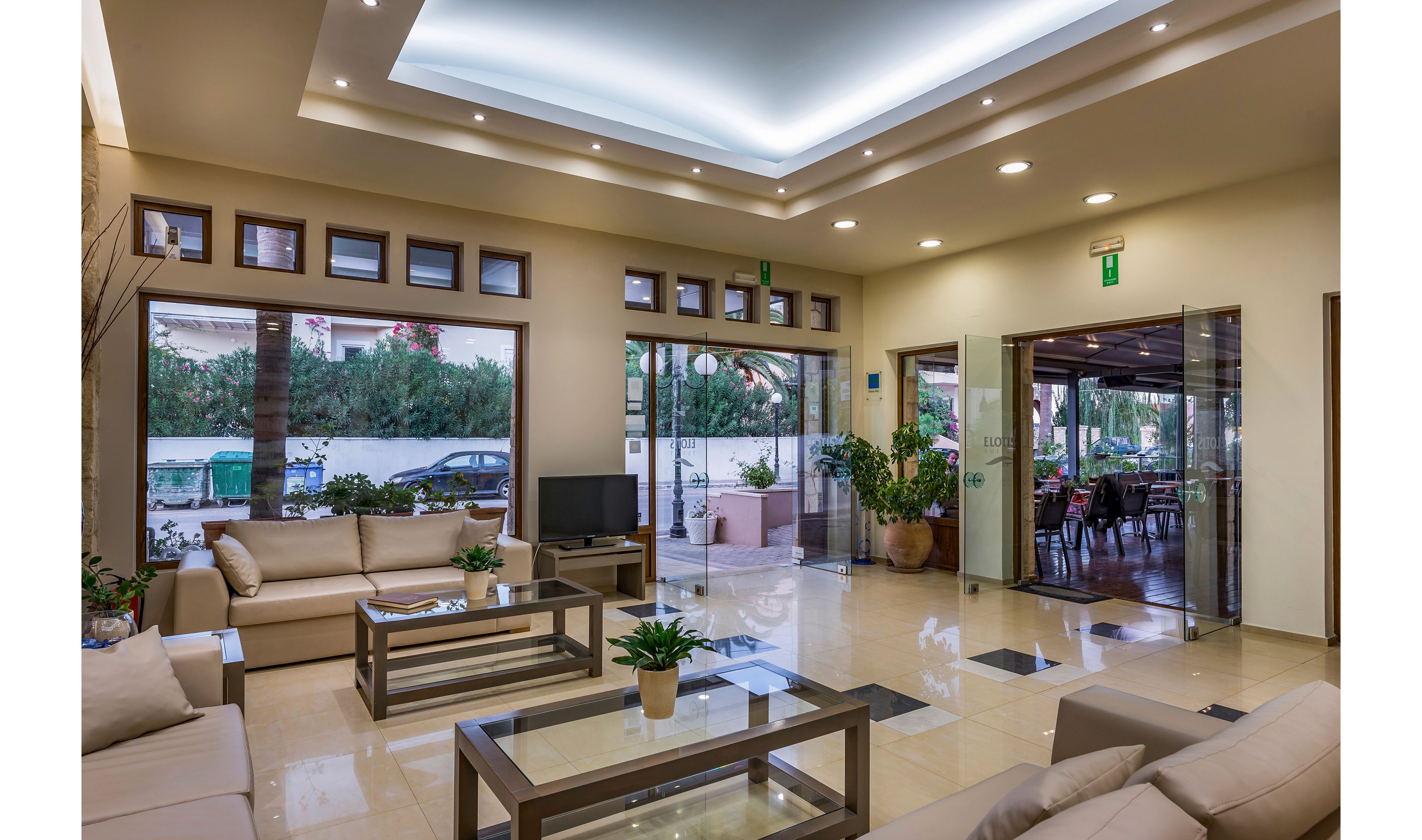 A contemporary lounge area features beige sofas and glass-topped coffee tables arranged on a polished tile floor. Large windows and sliding glass doors open to an outdoor patio with seating and potted plants. Recessed ceiling lights and a central illuminated panel create a bright, welcoming atmosphere. The space appears clean and organized, suitable for hotel guests or visitors to relax and socialize.
