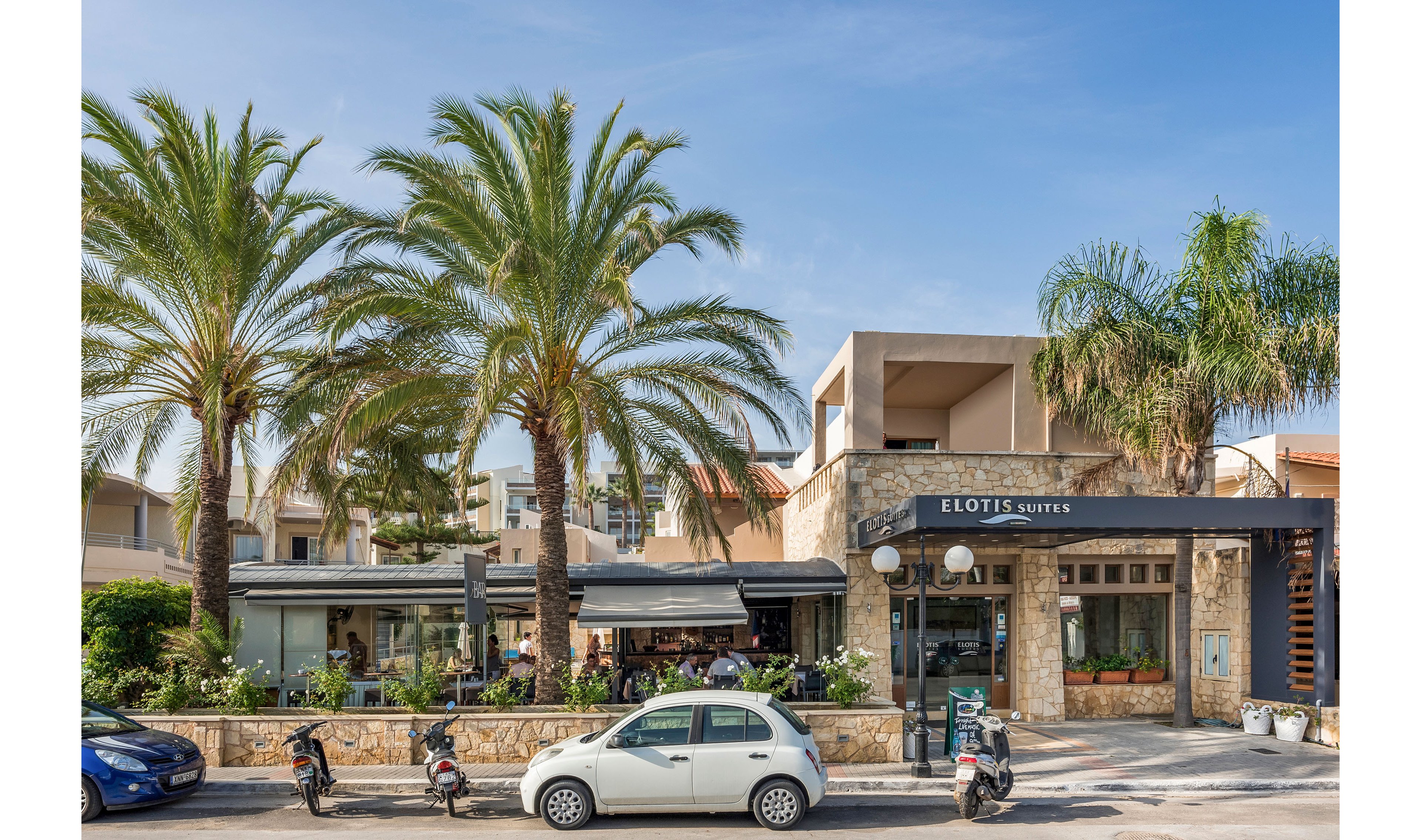 A Mediterranean hotel named Elotis Suites is shown from the street, framed by tall palm trees and a clear blue sky. The building features stone walls, balconies, and a shaded outdoor seating area where guests are gathered. Several scooters and a small car are parked along the road in front of the property, adding to the casual vacation atmosphere. The scene suggests a sunny resort destination with relaxed, open-air social spaces.