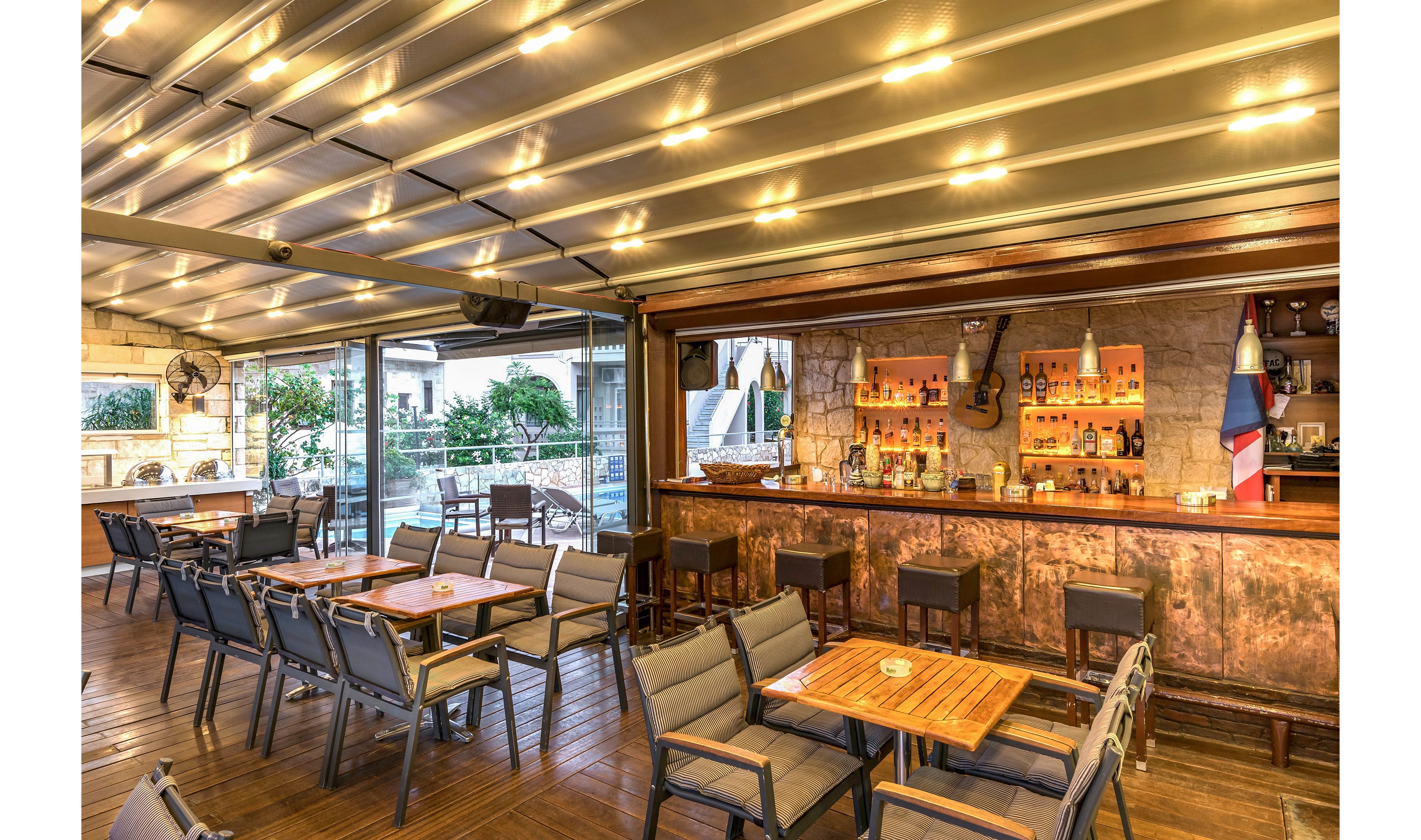 An inviting bar area with a warm wooden counter is set under a covered terrace. Multiple tables and cushioned chairs create a comfortable dining space with ample seating. Soft ceiling lights and decorative elements behind the bar add to the cozy atmosphere. The open sides of the terrace reveal greenery and nearby buildings, suggesting an outdoor urban setting.