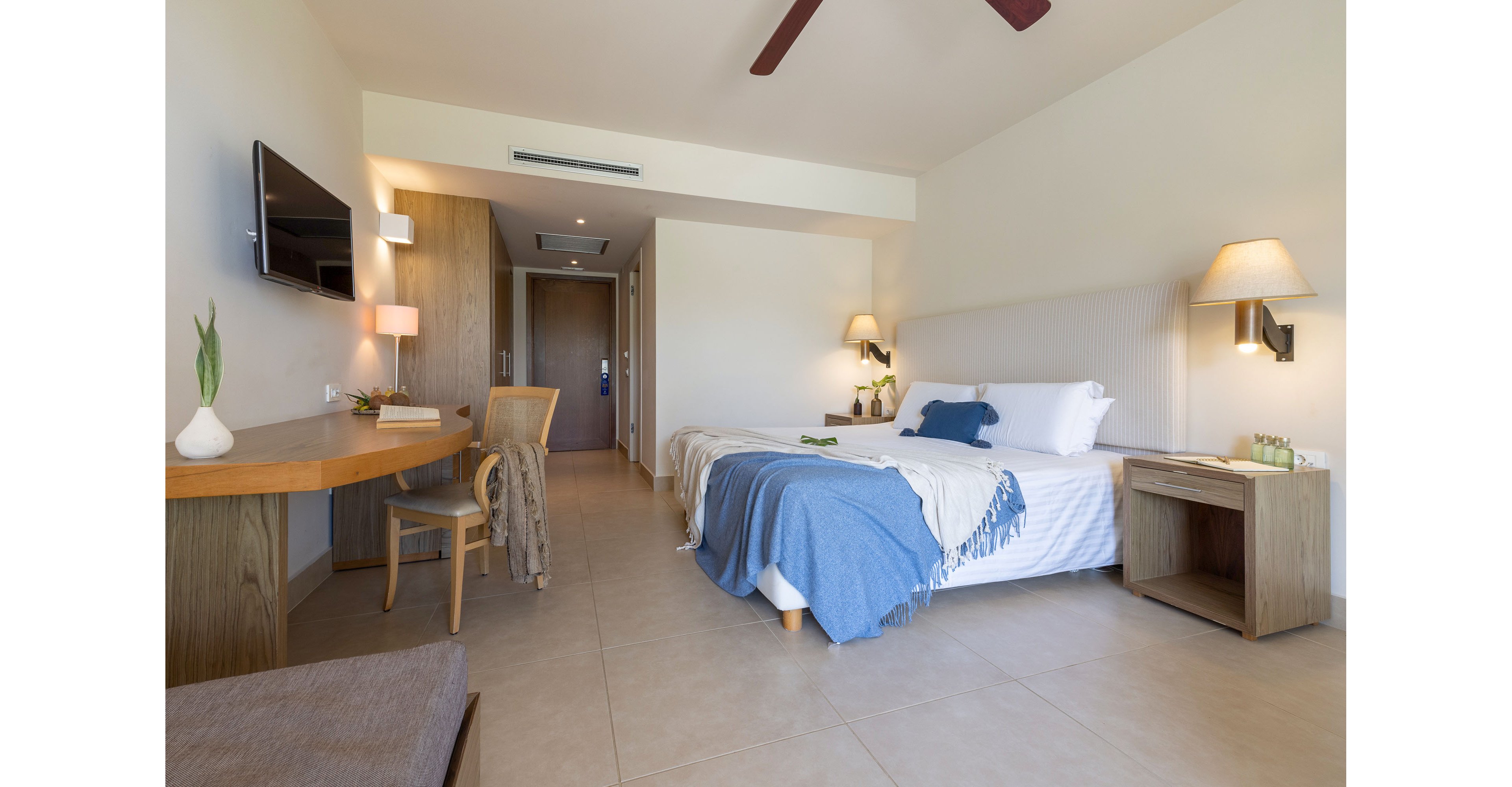 A contemporary hotel room features a neatly made bed with white linens and a blue throw blanket. A wooden desk with a chair and decorative items sits along one wall, opposite a wall-mounted television. Soft lighting from wall sconces and neutral-colored tile flooring create a calm, inviting atmosphere. The room extends toward an open doorway and hallway, emphasizing spaciousness and clean design.