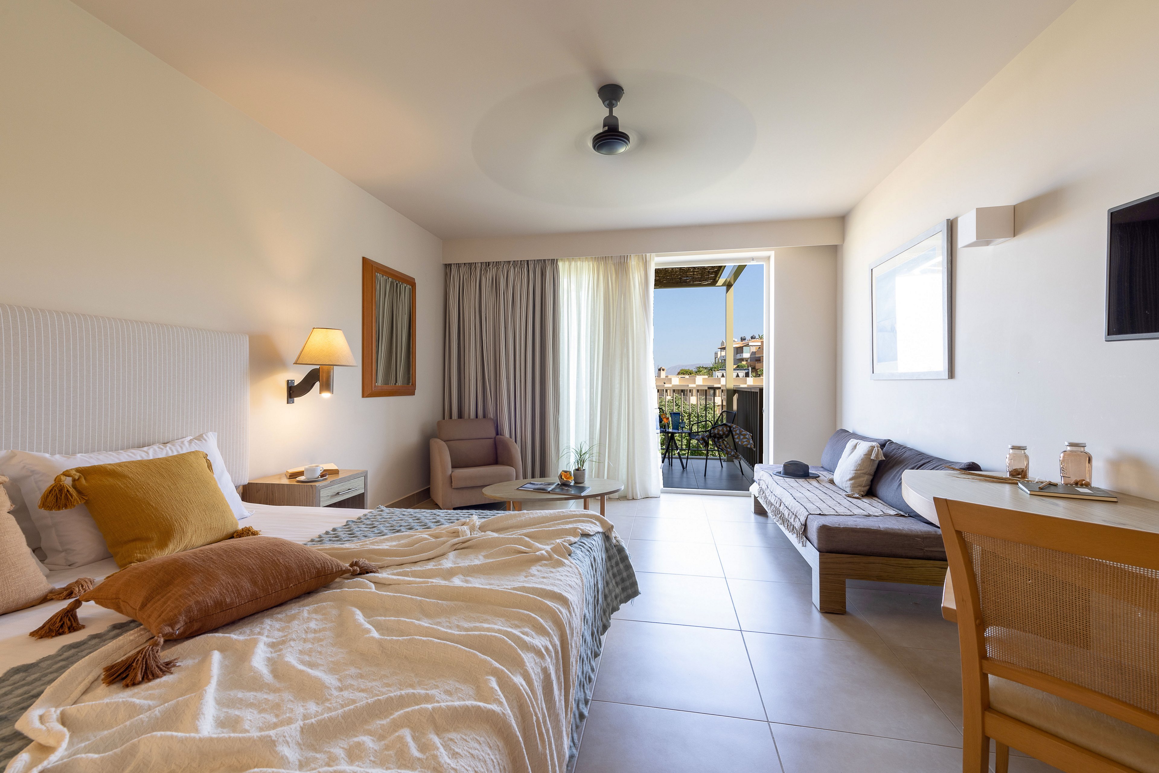 A sunlit hotel bedroom features an unmade double bed with cozy pillows and throws. The room includes a lounge chair, side table, and a daybed along the opposite wall. Neutral tones and soft lighting create a calm, inviting atmosphere. Large glass doors open to a balcony overlooking a scenic outdoor landscape.