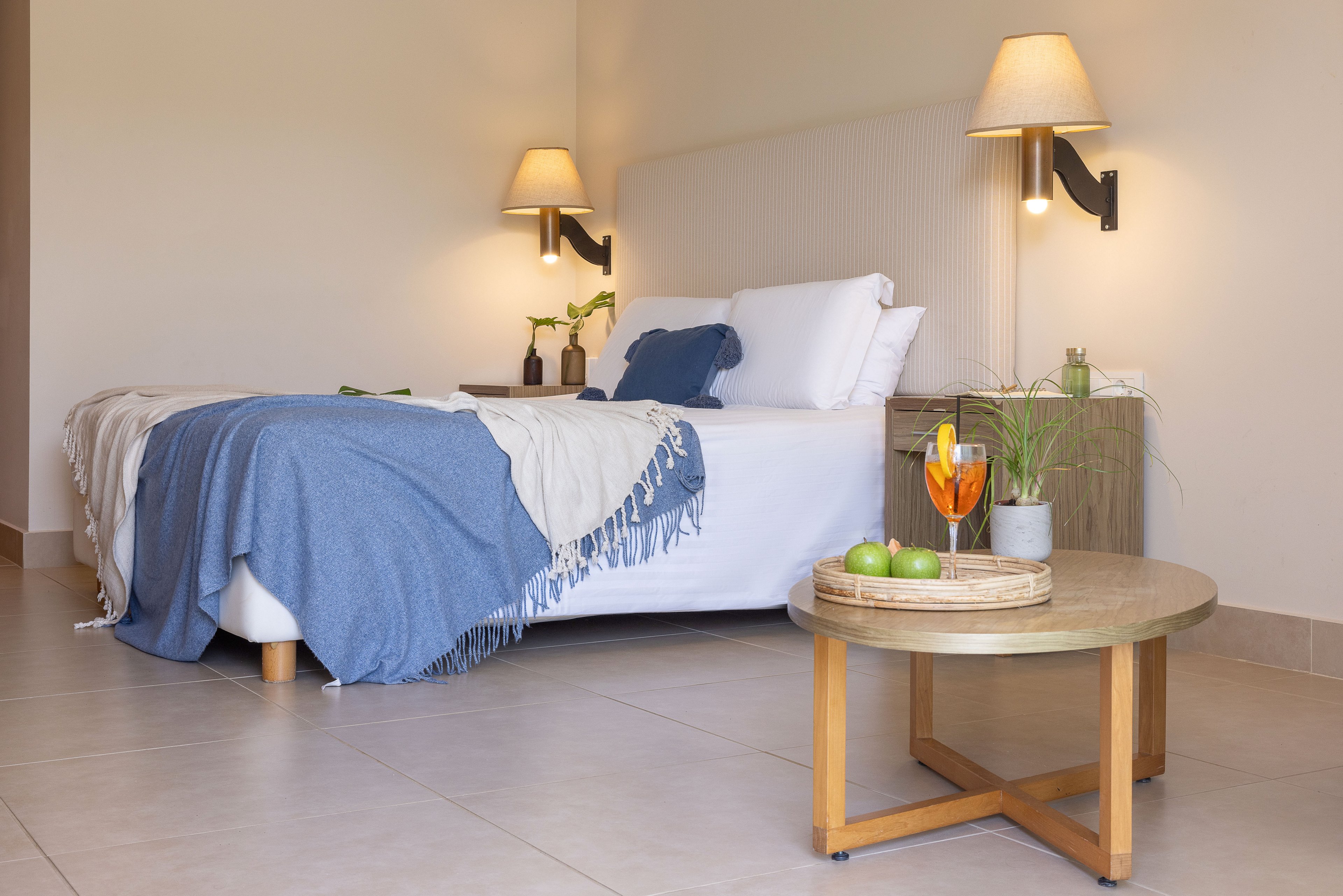 A neatly made bed with white linens and a blue fringed throw blanket is the focal point of this modern bedroom. The room features light-colored walls, tiled flooring, and simple wooden furniture including a bedside table and a round coffee table. Wall-mounted lamps provide warm ambient lighting, while green plants and a citrus drink with limes add fresh, colorful accents. The overall setting feels calm, minimal, and inviting, suitable for a contemporary home or hotel interior.