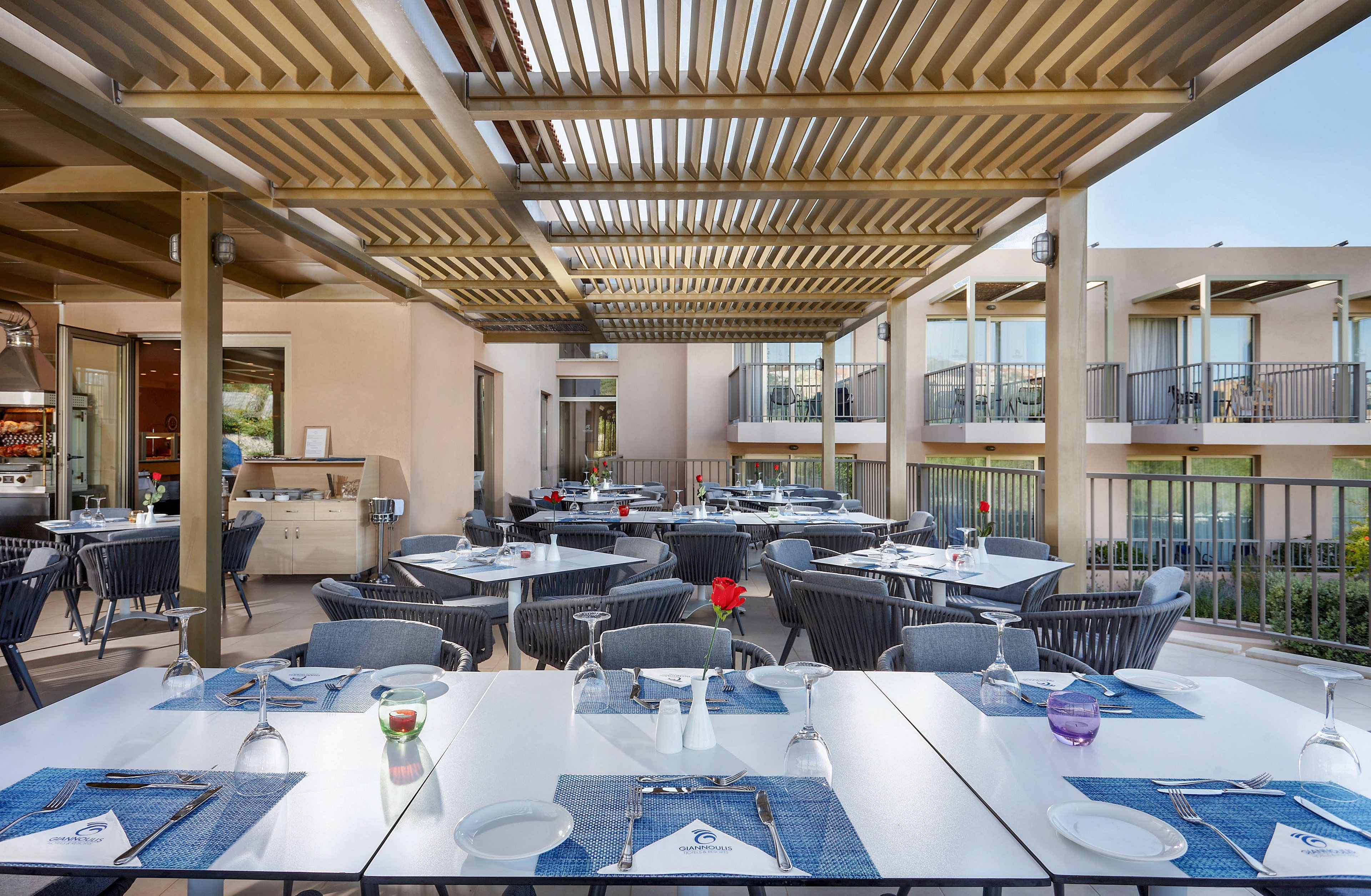 An open-air restaurant terrace features neatly arranged dining tables under a modern slatted pergola. Tables are set with plates, glasses, cutlery, and folded napkins on blue placemats, ready for guests. Contemporary chairs surround the tables, and a single red flower decorates one of them, adding a touch of color. In the background, a multi-story building with balconies overlooks the terrace, suggesting a hotel or resort setting.