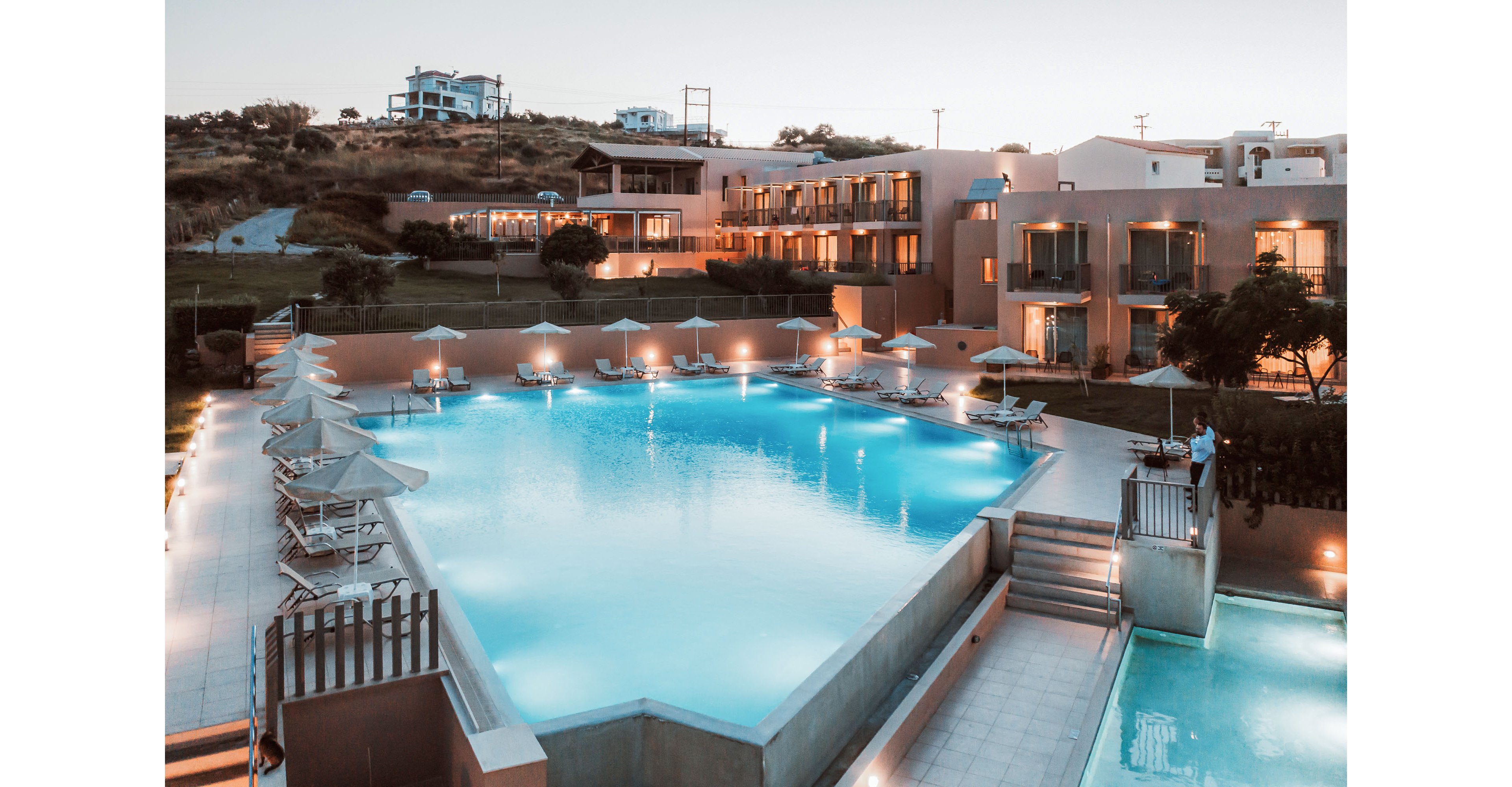 A large outdoor swimming pool is the central focus of a modern resort complex at dusk. Multi-story hotel buildings with balconies surround the pool area, with sun loungers and umbrellas neatly arranged along the deck. Soft artificial lighting reflects on the calm water, creating a tranquil atmosphere. The scene suggests a relaxing vacation setting in a warm climate.