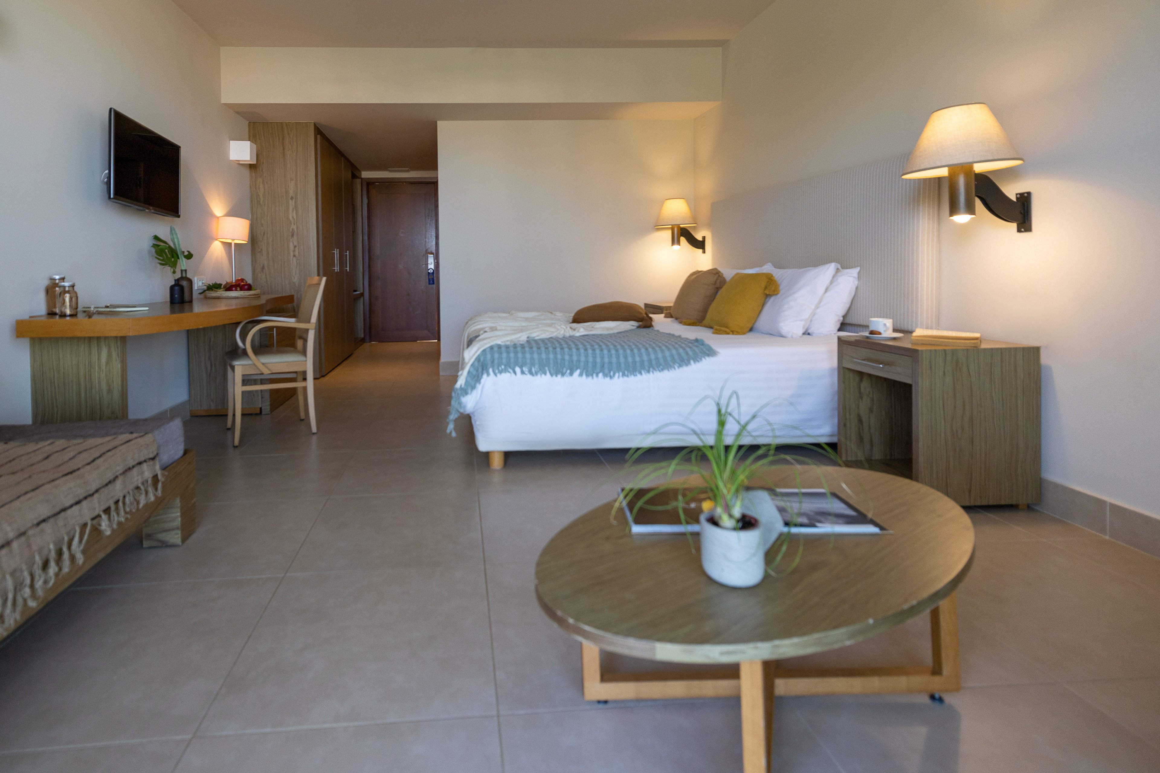A contemporary hotel room features a neatly made double bed with white linens and accent pillows. The space includes a wooden desk with a chair, wall-mounted television, and bedside tables with warm wall lamps. A low round coffee table with a small potted plant and magazines sits in the foreground on a tiled floor. Neutral colors and simple furnishings create a calm, minimalist atmosphere suitable for relaxation or work.