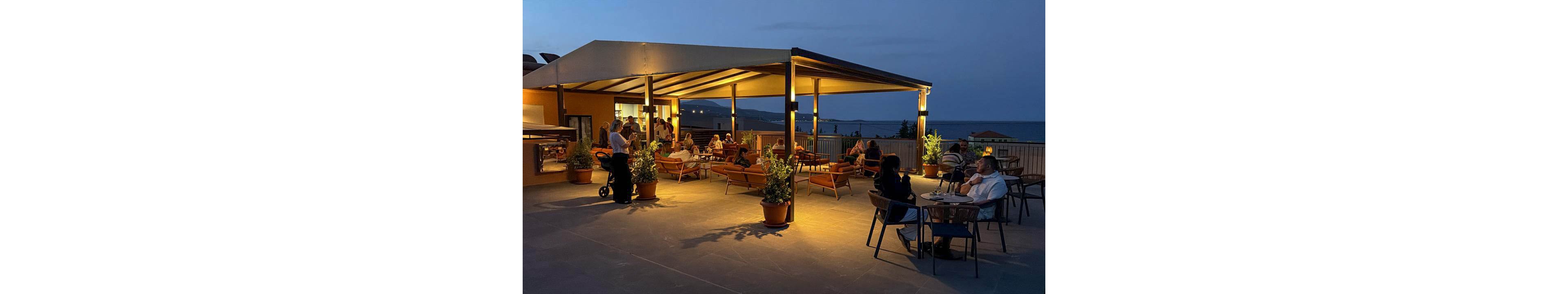 An open-air rooftop terrace restaurant is softly lit in the evening, with warm lights glowing under a modern canopy. Groups of people sit at tables and lounge chairs, talking and enjoying drinks in a relaxed atmosphere. Potted plants decorate the spacious patio, adding greenery to the contemporary setting. In the background, the sea and distant hills are visible under the darkening blue sky.