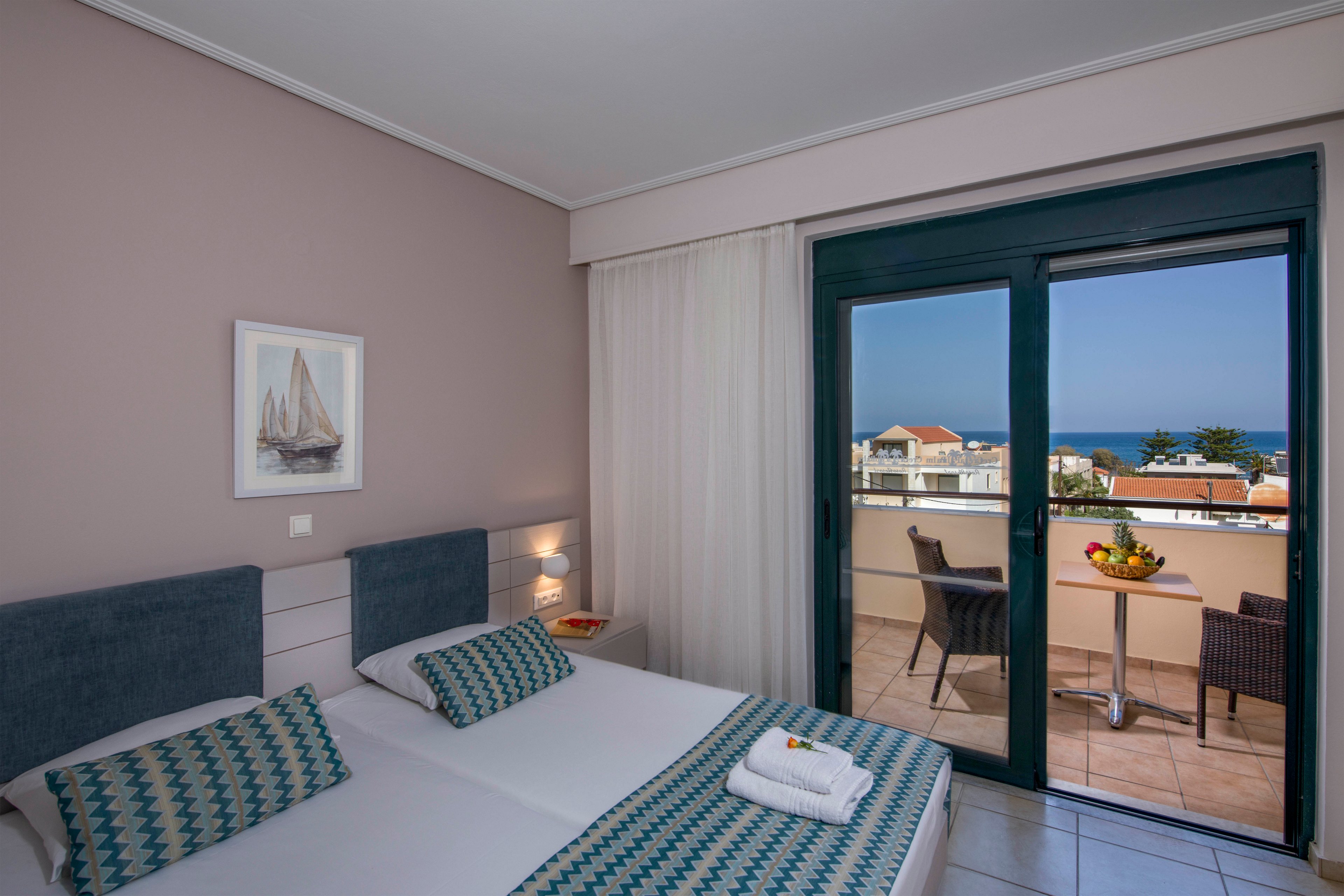A tidy hotel bedroom features two single beds pushed together with patterned pillows and a folded towel on top. Soft pastel walls and a framed sailboat picture create a calm, coastal atmosphere. Large glass doors open onto a tiled balcony with a small table, two wicker chairs, and a bowl of fruit. Beyond the balcony, rooftops and the distant sea are visible under a clear sky.