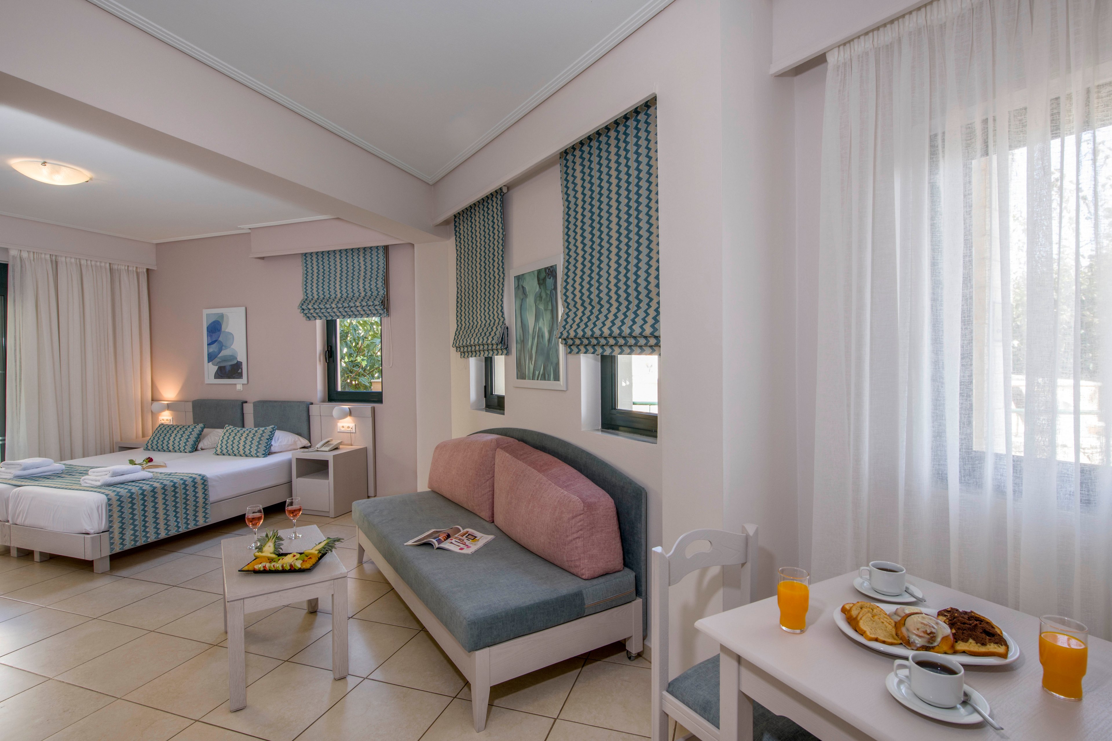 A spacious hotel room features a neatly made double bed with striped bedding and a small bedside table. A sofa with cushions sits against the wall beneath several windows with patterned teal curtains. In the foreground, a small table holds plates of pastries, a cup and saucer, and glasses of orange juice. Soft natural light filters through sheer white curtains, creating a calm and inviting atmosphere. The room is decorated in light pastel tones with tiled flooring and simple wall art.