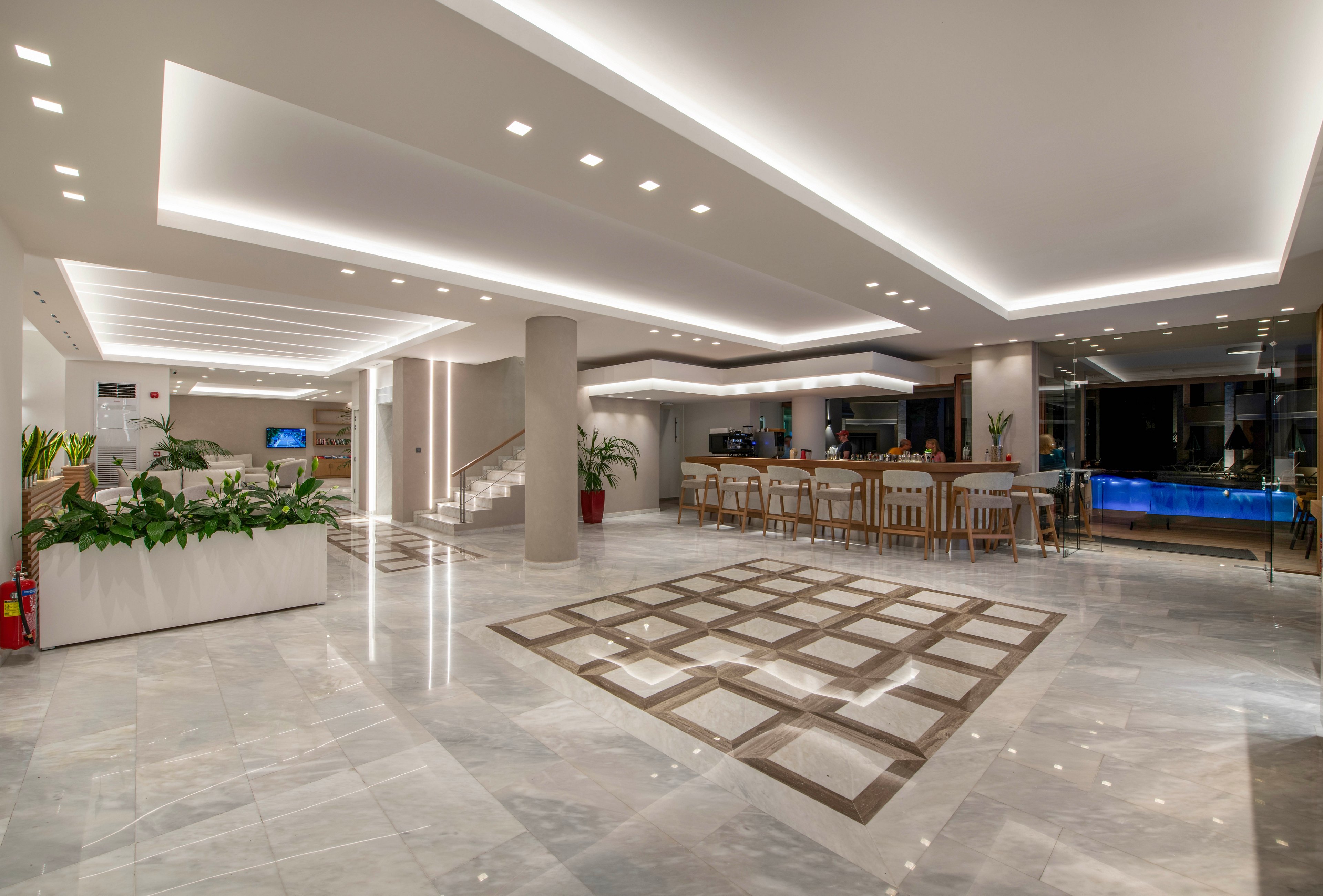 A spacious, contemporary hotel lobby features polished marble floors and recessed ceiling lighting. A seating and bar area with high stools is visible on the right, adjacent to a glass wall and illuminated blue accent feature. On the left, indoor plants and a staircase add greenery and vertical interest to the clean architectural lines. The overall scene is bright, minimal, and welcoming, emphasizing modern interior design.