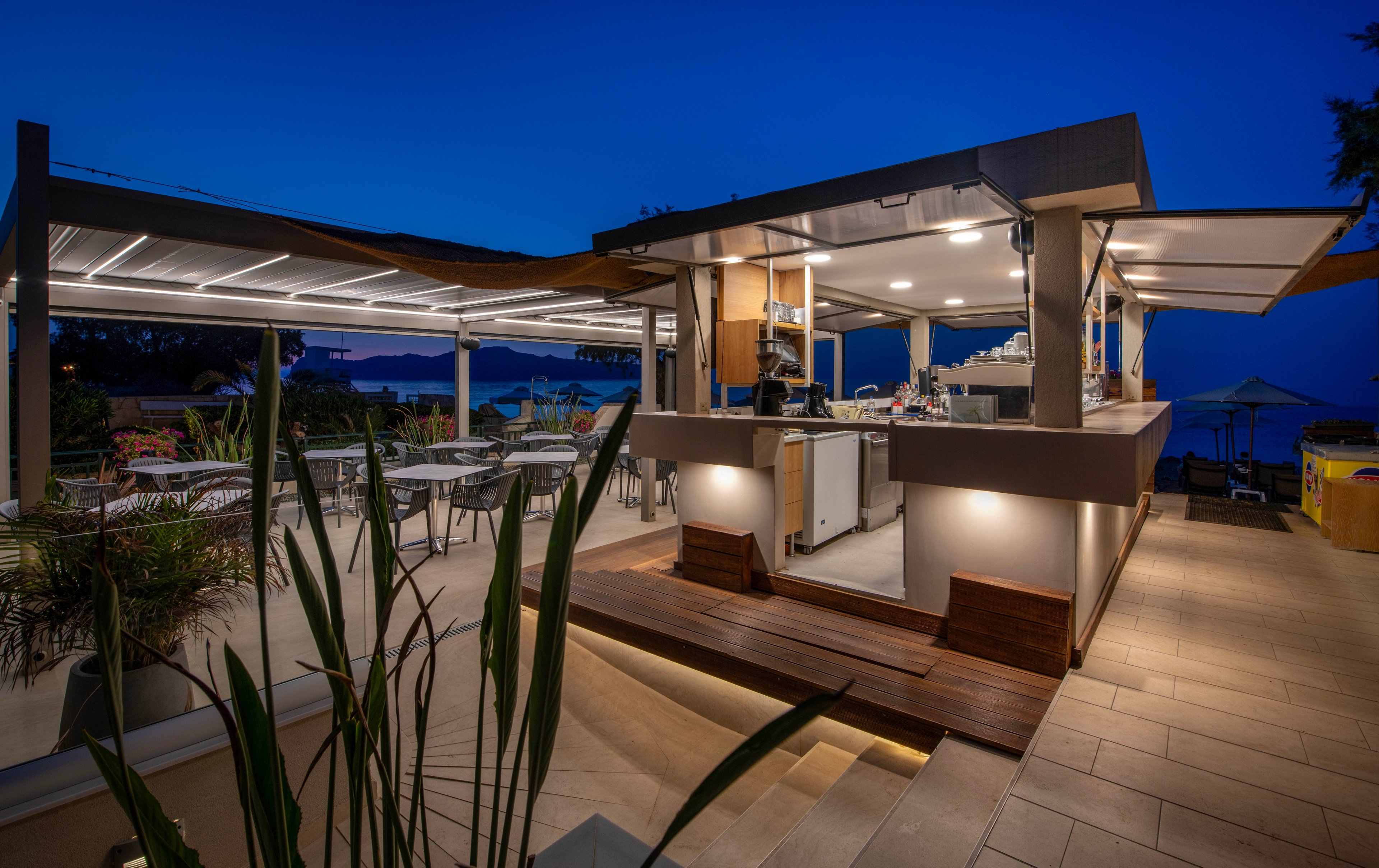 A contemporary open-air bar is illuminated with warm lights during blue hour. The structure features clean lines, a wooden deck, and integrated lighting along the steps and counters. Empty tables and chairs are arranged under a covered terrace, overlooking a distant sea and coastline. Lush plants frame the foreground, enhancing the relaxed, upscale atmosphere of the evening setting.