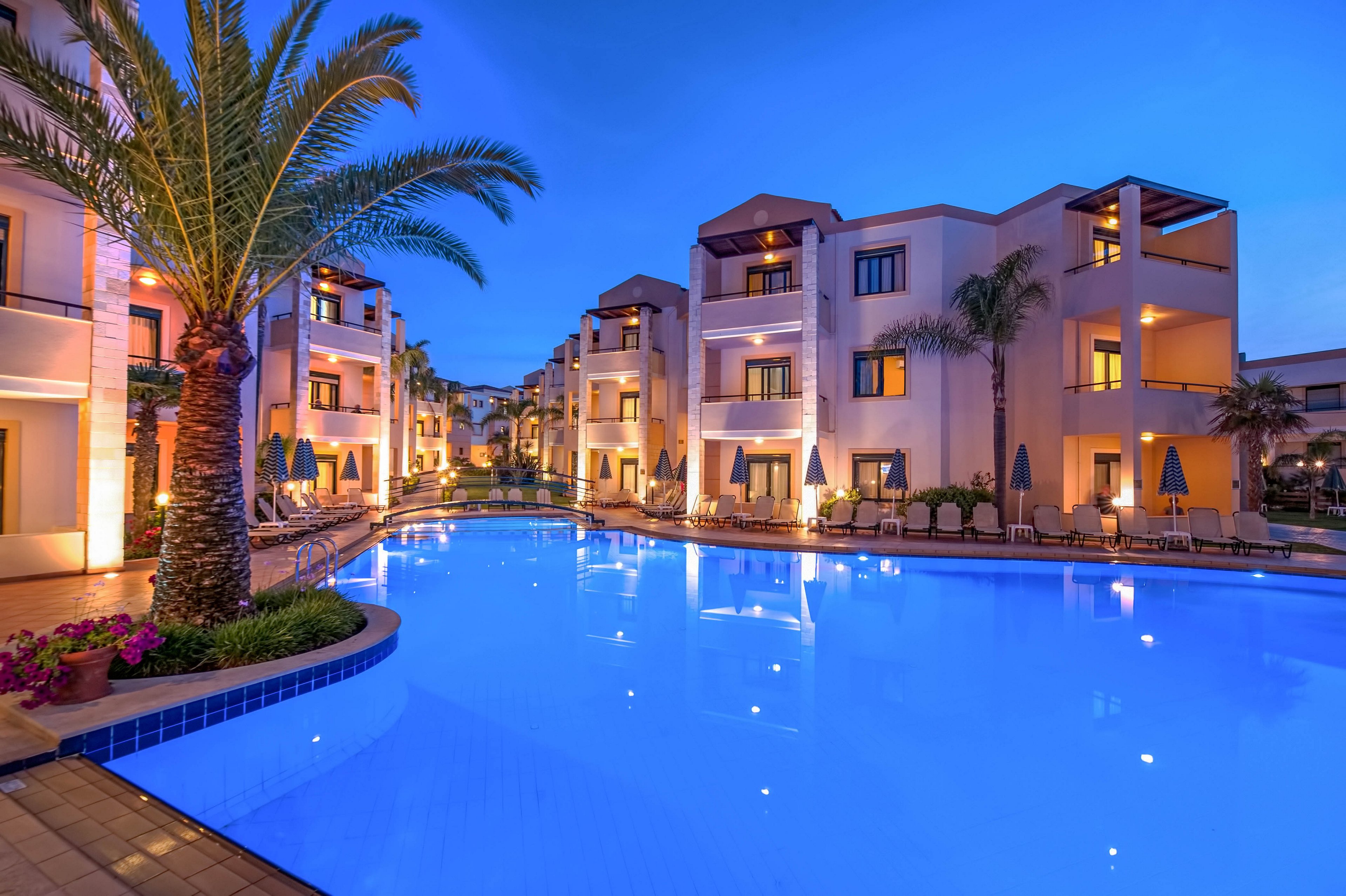 A large illuminated swimming pool is surrounded by modern resort-style apartment buildings at dusk. Palm trees and poolside lounge chairs line the water, creating a relaxing vacation atmosphere. Warm lights from balconies and windows contrast with the deep blue evening sky and glowing pool. The scene suggests a tranquil holiday destination with comfortable outdoor amenities.