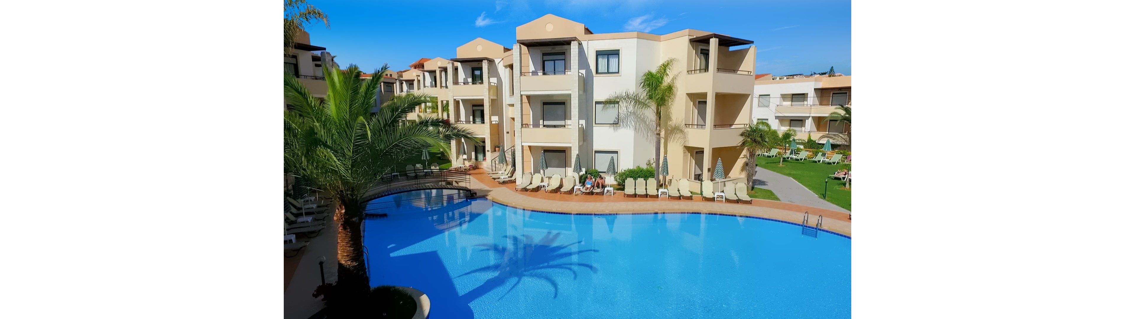A large curved swimming pool sits in front of a modern resort-style apartment complex. Cream-colored buildings with balconies and terraces surround the pool area, with sun loungers neatly arranged along the water’s edge. Palm trees and manicured lawns add a tropical feel to the scene. A few people relax on the loungers, suggesting a calm vacation atmosphere under a clear blue sky.