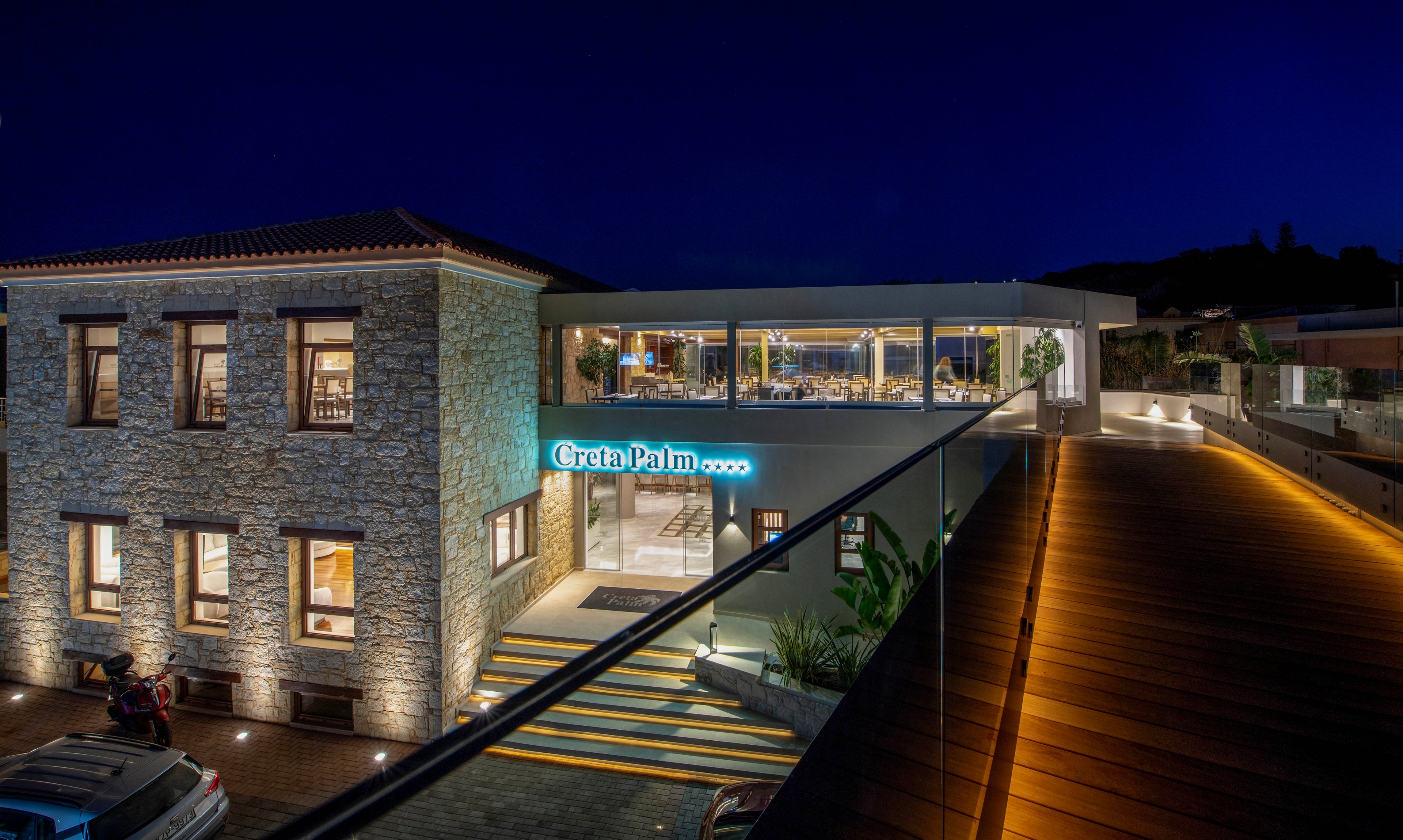 A contemporary hotel complex is shown at night with warm interior lights glowing through large windows. The main stone building stands beside a modern glass-fronted structure connected by an elevated walkway. A neon sign reading "Creta Palm" is prominently displayed above the entrance. The scene features clean architectural lines, landscaped plants, and subtle pathway lighting against a deep blue sky.