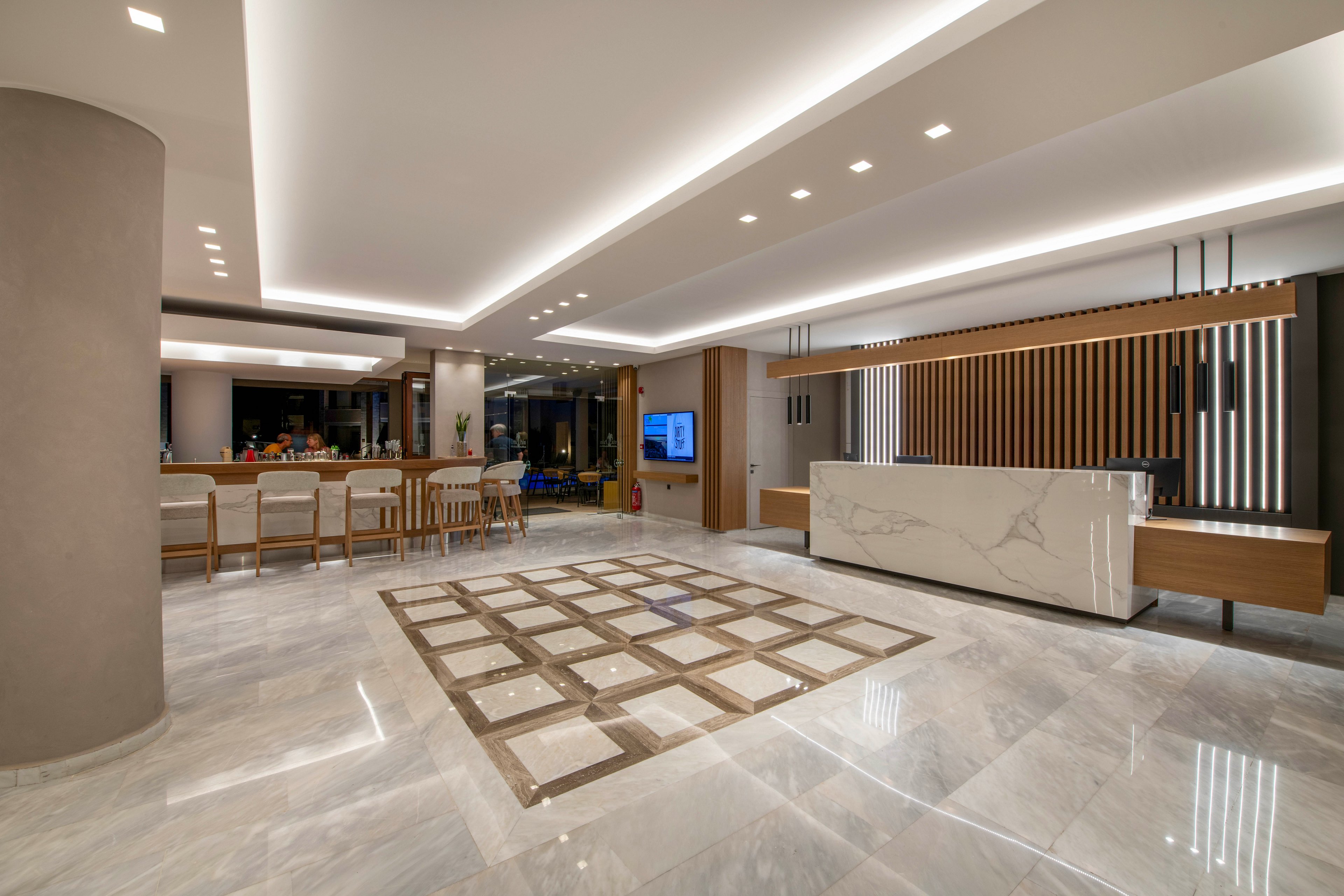 Spacious contemporary lobby interior featuring a sleek marble reception desk and polished stone flooring. A geometric tile pattern decorates the center of the floor, adding visual interest to the open space. Warm recessed ceiling lights and cove lighting create a soft, inviting ambiance. In the background, a bar area with high stools and large windows suggests a hotel or upscale hospitality setting.