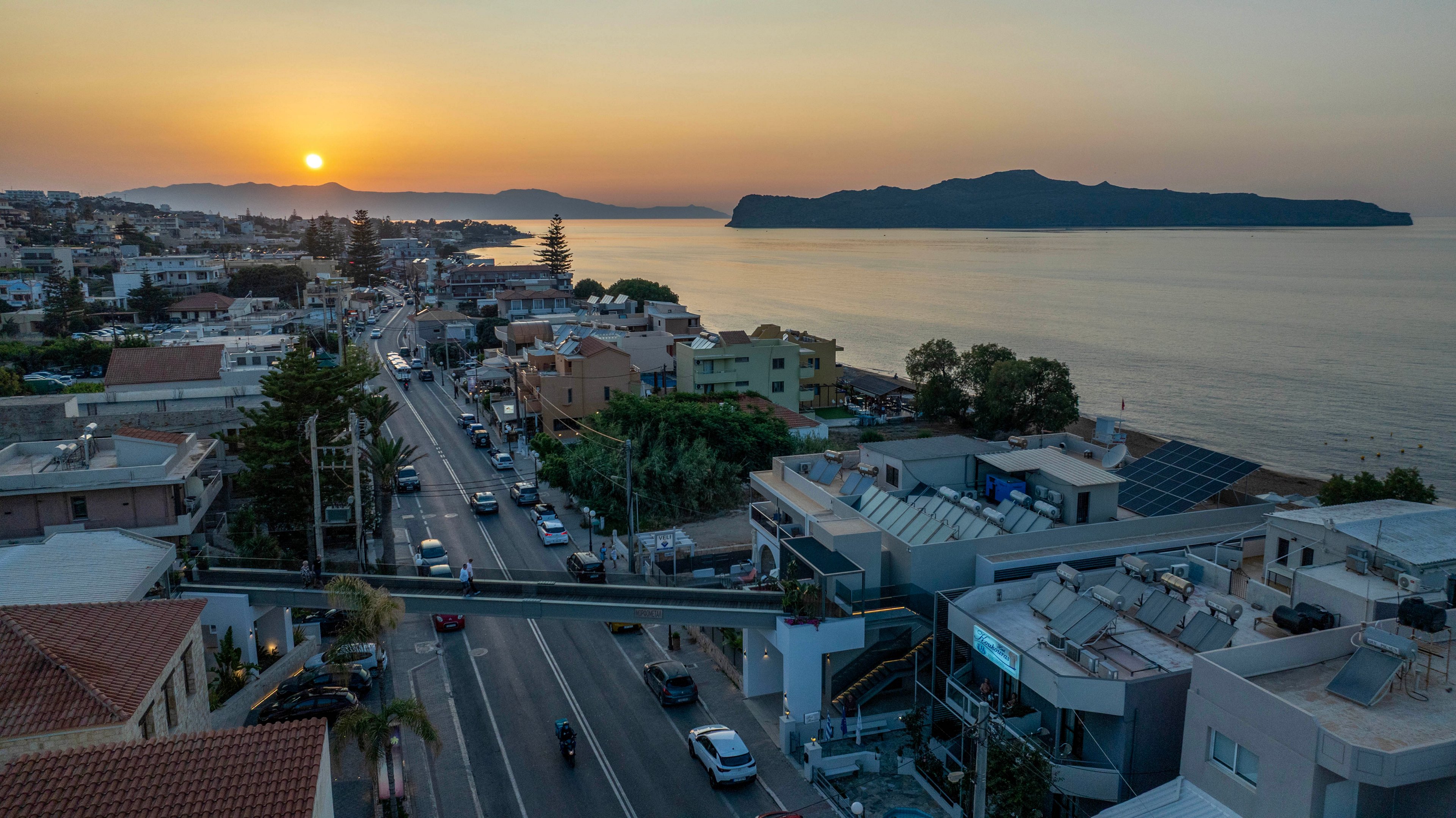 An elevated view captures a coastal town at sunset, with the sun low over the horizon and reflecting on the calm sea. A main road runs parallel to the shoreline, lined with buildings, shops, and houses, while cars travel in both directions. Rooftops with terraces and solar panels are visible in the foreground, and a long island or peninsula stretches across the distant water. The warm evening light creates a tranquil atmosphere over the seaside community.