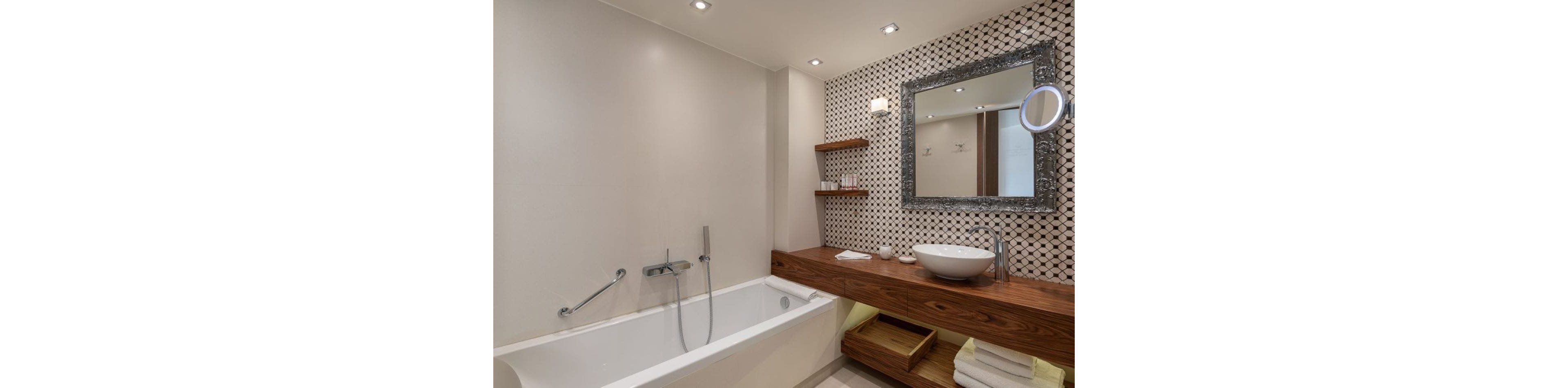 Contemporary bathroom interior featuring a white bathtub with a wall-mounted faucet and handheld shower. A long wooden vanity with a vessel sink and chrome faucet runs along one side, complemented by open shelves holding glassware and accessories. The wall behind the vanity is covered with geometric patterned tiles, and a large framed mirror with an integrated round magnifying mirror adds visual interest. Soft recessed lighting and neatly folded white towels create a clean, spa-like atmosphere.