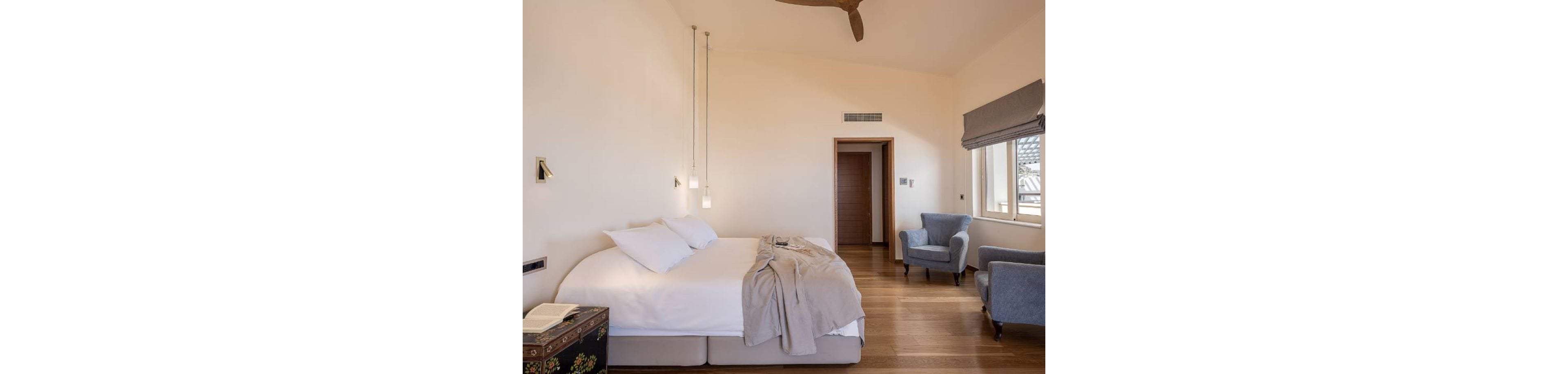 A bright, minimalist bedroom features a large bed with white linens and a light gray throw blanket. Two pendant lights hang above the bedside area, illuminating a small chest with an open book. The room has warm wooden flooring, neutral walls, and a doorway leading to another space. A pair of upholstered armchairs sit near a window with gray Roman shades, adding comfort and relaxation to the setting.