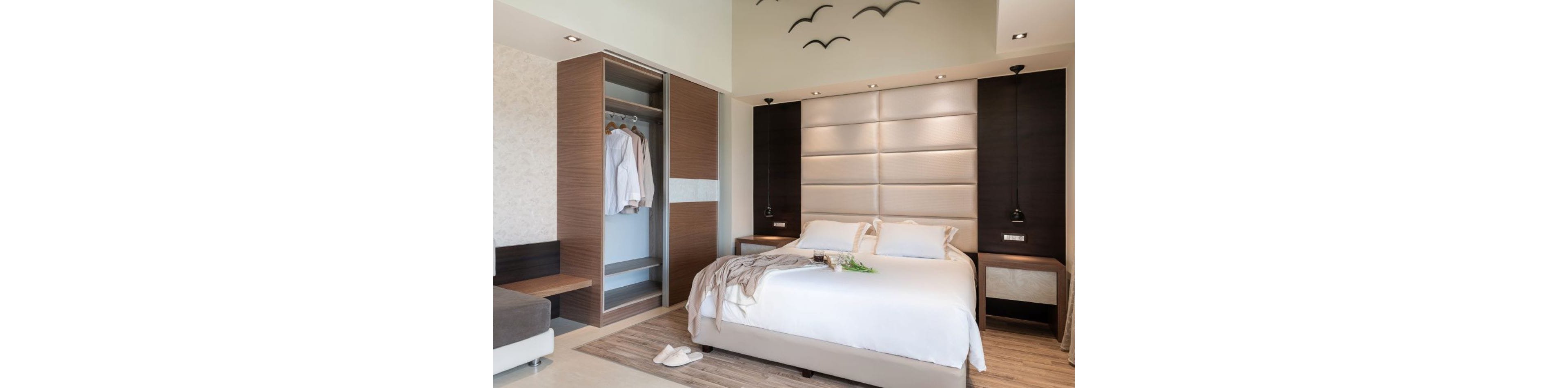 A contemporary bedroom features a large double bed with white linens and soft beige upholstery on the headboard. An open wardrobe with wooden doors reveals neatly hung white clothing on hangers. Minimalist bedside tables with pendant lights flank the bed, while slippers and a draped throw add a lived-in touch. Decorative bird silhouettes on the wall and neutral tones create a calm, inviting atmosphere.