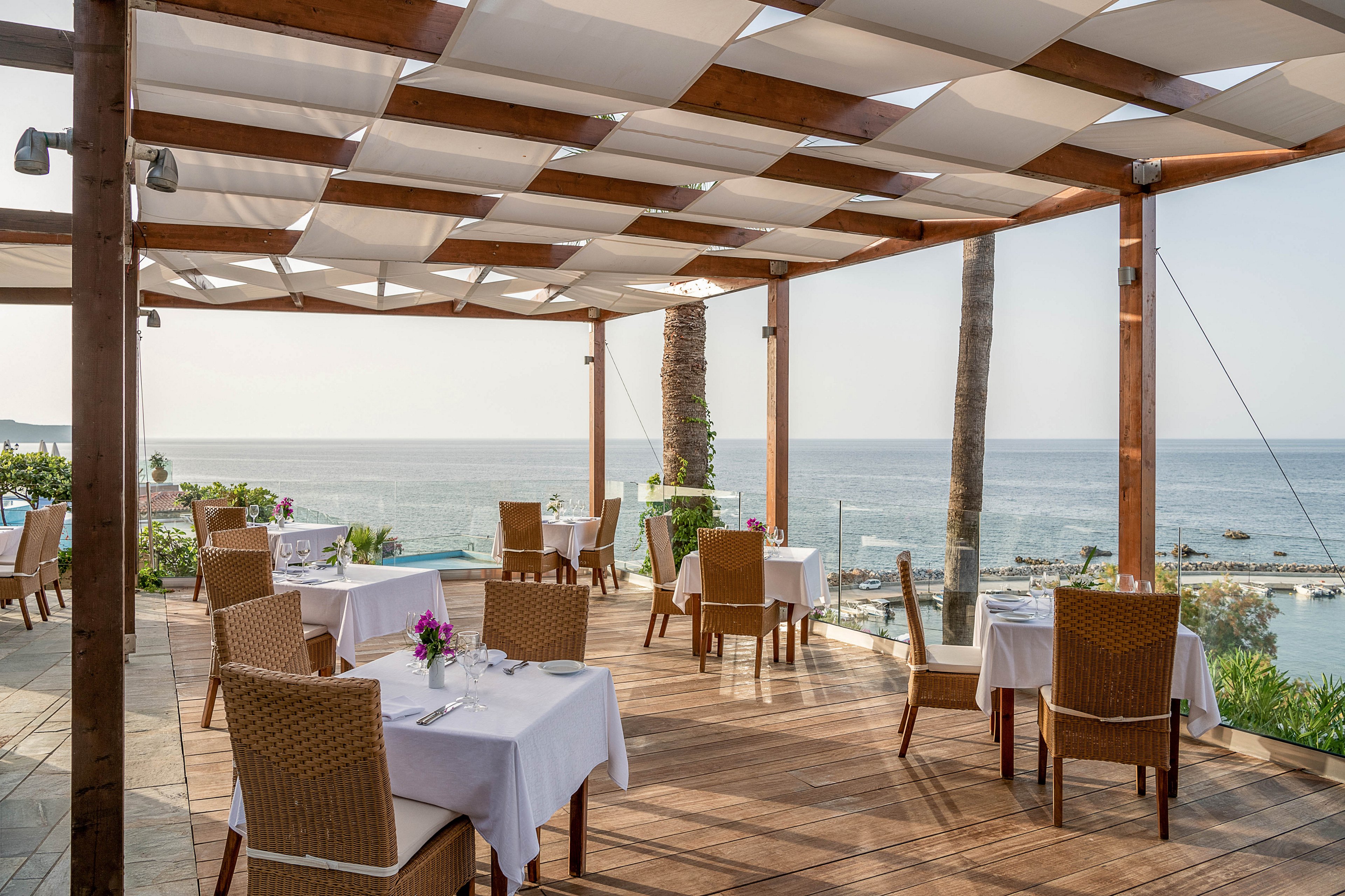 An elegant open-air restaurant terrace overlooks a calm ocean. Wooden decking and a modern pergola frame neatly arranged tables covered with white linens. Wicker chairs surround each table, some decorated with small floral centerpieces. The bright daylight and clear sky create a relaxed, upscale coastal dining atmosphere.