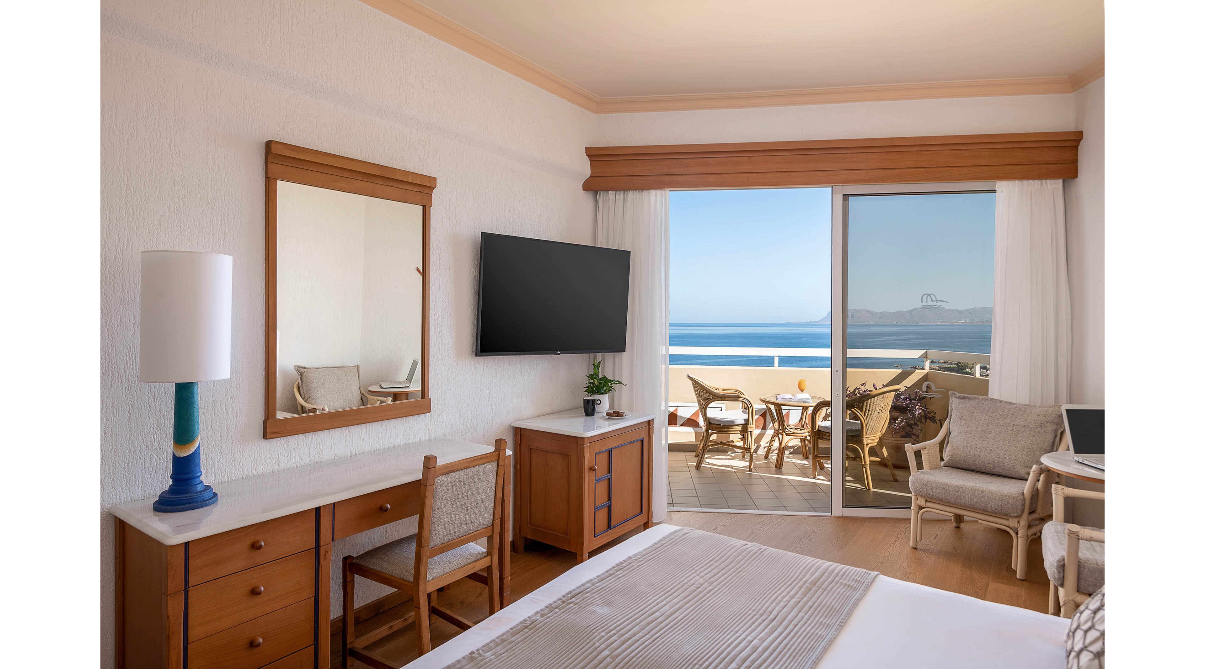 A bright hotel room features a neatly made bed, wooden furniture, and a wall-mounted flat-screen television. Large sliding glass doors open onto a balcony with chairs and a table overlooking a calm ocean and distant coastline. Natural light fills the space, highlighting neutral tones and simple decor. The scene suggests a relaxing seaside vacation setting with indoor comfort and outdoor leisure combined.