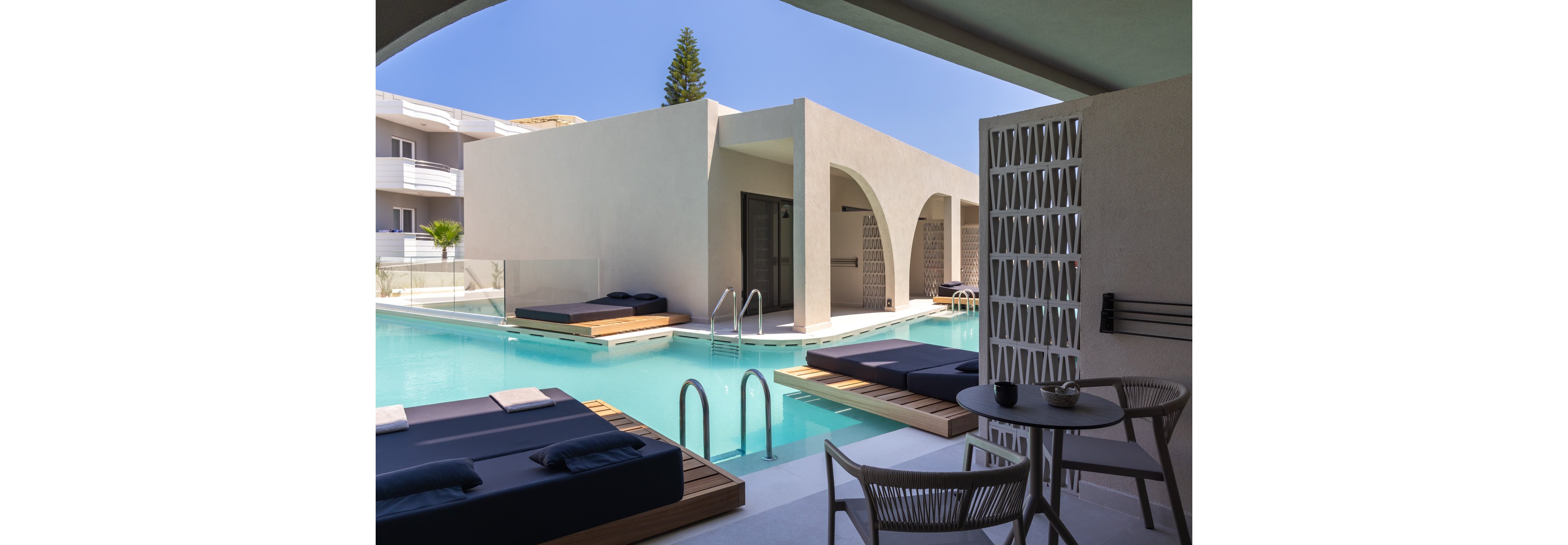 Contemporary poolside cabanas sit beside a clear blue swimming pool in a resort setting. Minimalist architecture with clean lines, arches, and light-colored walls creates a luxurious atmosphere. Dark cushioned daybeds and a small table with chairs provide comfortable seating overlooking the water. The scene is calm and inviting, suggesting a relaxing vacation or leisure experience under a bright sky.