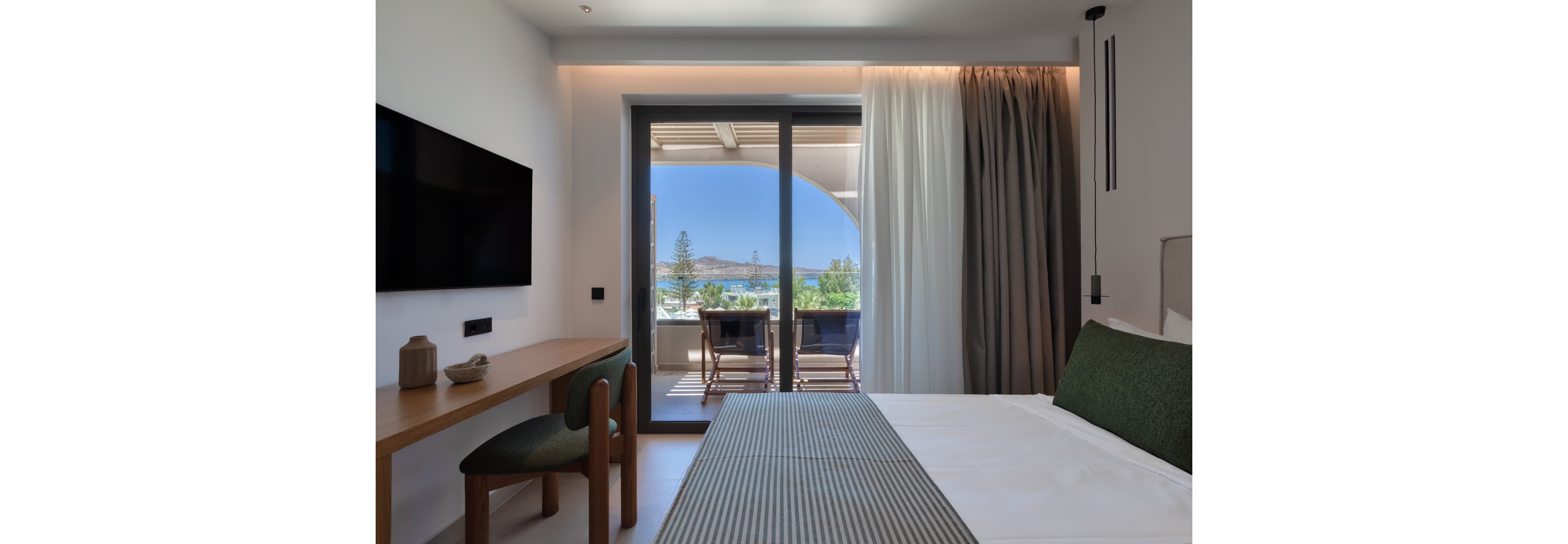 A contemporary hotel room features a neatly made bed with green accents and a wooden desk with a chair. Large sliding glass doors open to a balcony with two chairs and a table, overlooking a scenic coastal landscape. Soft natural light fills the space, complemented by sheer and blackout curtains and minimalist decor. A wall-mounted television and subtle lighting create a calm, relaxing atmosphere ideal for vacation or business travel.