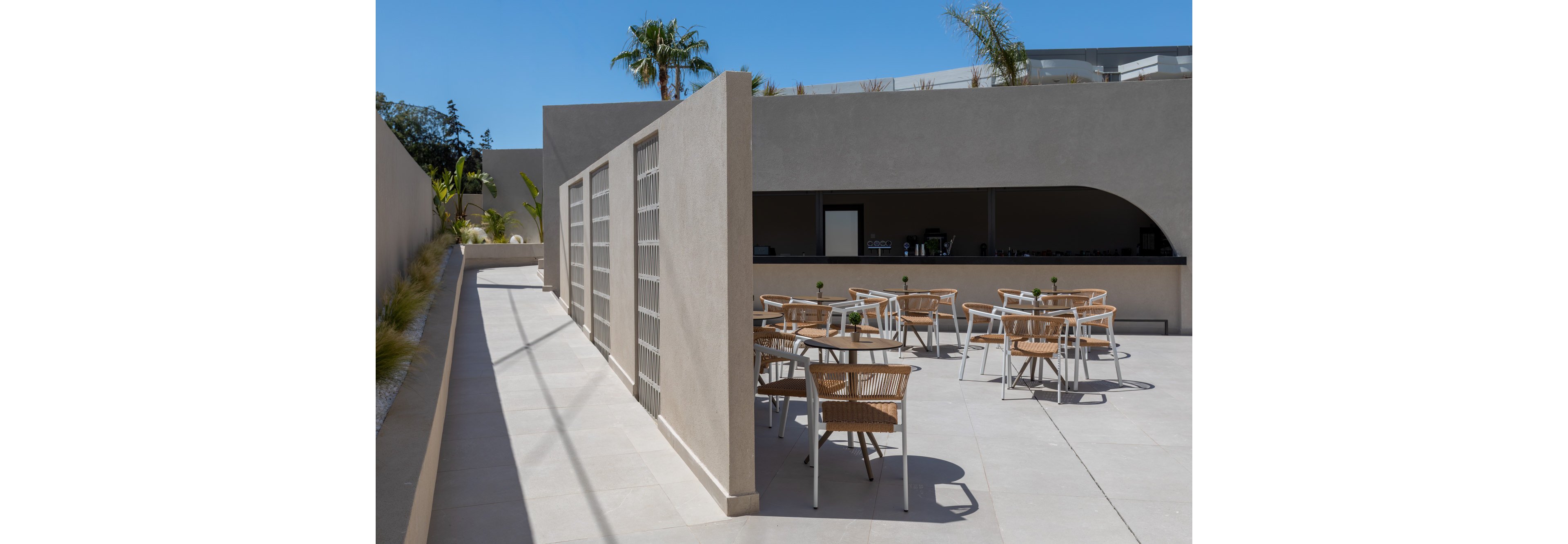 A contemporary outdoor cafe terrace features light wooden chairs and round tables arranged on a smooth stone patio. The setting is defined by clean beige concrete walls and an open bar area with a large curved opening. Small plants on the tables add a touch of greenery, while palm trees rise above the structure in the background. Bright sunlight and sharp shadows emphasize the minimalist, modern architectural style.