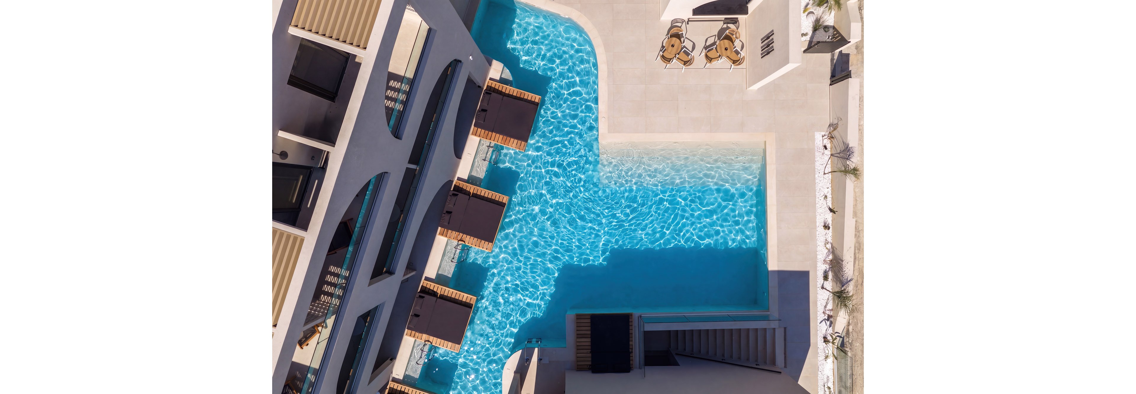 A bright turquoise swimming pool is captured from above in a contemporary resort setting. Sun loungers line one side of the pool beside a modern building with geometric architectural details. On the surrounding terrace, a small table with chairs sits in the sun, and stairs lead down to another level. The scene conveys a clean, minimalist, and luxurious vacation atmosphere.