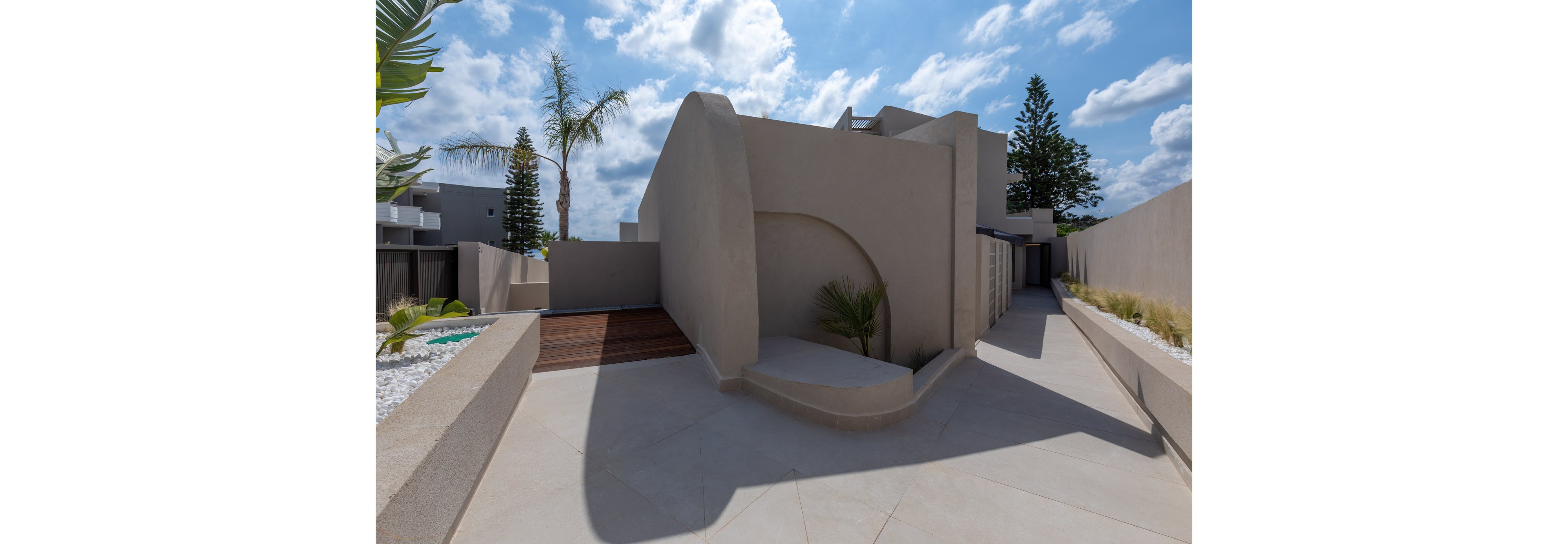 A contemporary residential courtyard features smooth, curved concrete walls and a clean minimalist design. Light-colored paving leads toward the entrance, framed by geometric forms and a small planted area with palm trees. The setting is outdoors under a bright blue sky with scattered clouds, creating strong shadows and contrast. Subtle landscaping with gravel beds and low greenery enhances the modern architectural aesthetic.