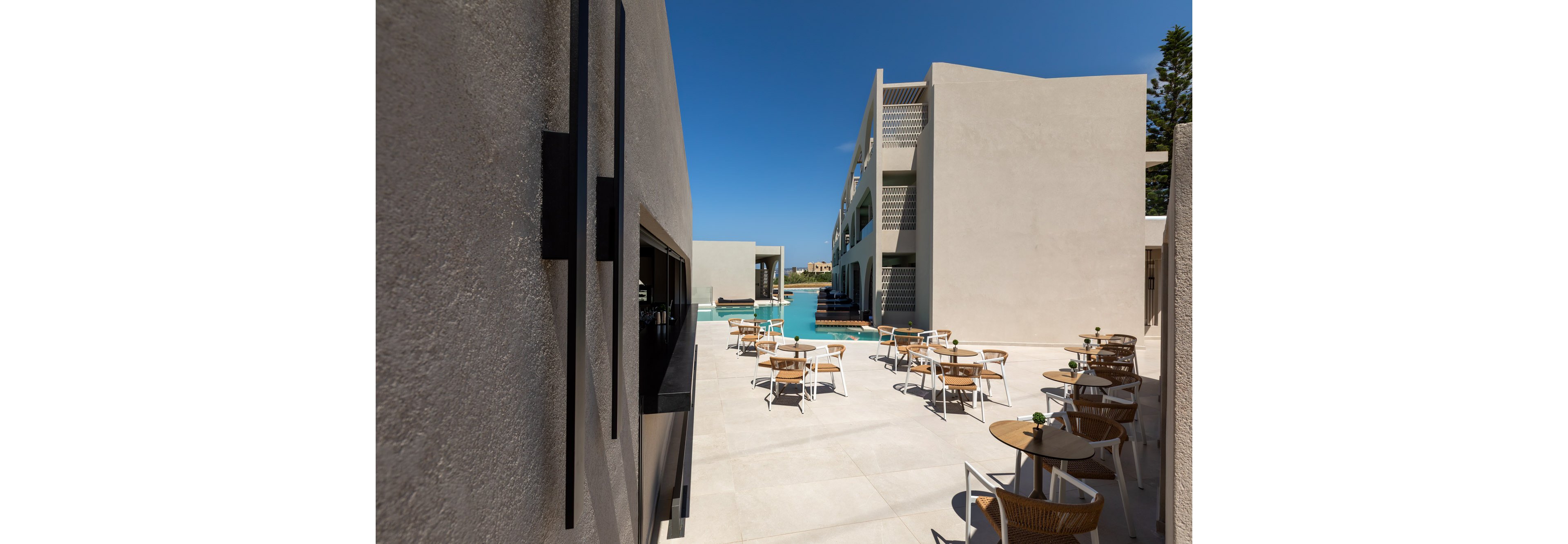 A contemporary hotel terrace features neatly arranged tables and chairs under a clear blue sky. The open-air seating area is framed by minimalist concrete buildings and overlooks a long swimming pool. Sunlight casts sharp shadows across the pale tiled floor, emphasizing the clean architectural lines. The scene suggests a relaxed, resort-style atmosphere ideal for outdoor dining or lounging.