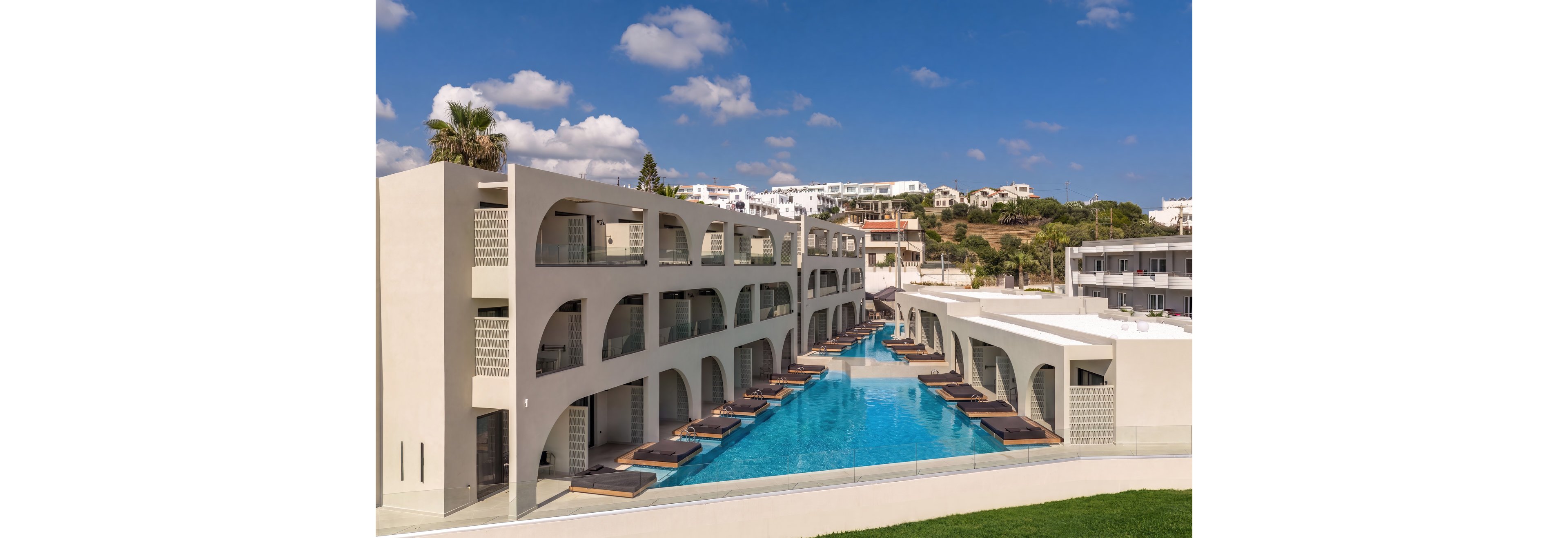 Contemporary low-rise resort buildings frame a long outdoor swimming pool lined with sun loungers and umbrellas. The architecture features clean lines, arches, and neutral tones that contrast with the bright blue water. In the background, white hillside houses and greenery suggest a coastal or Mediterranean setting under a clear, partly cloudy sky. The scene conveys a relaxed vacation atmosphere focused on leisure and poolside comfort.