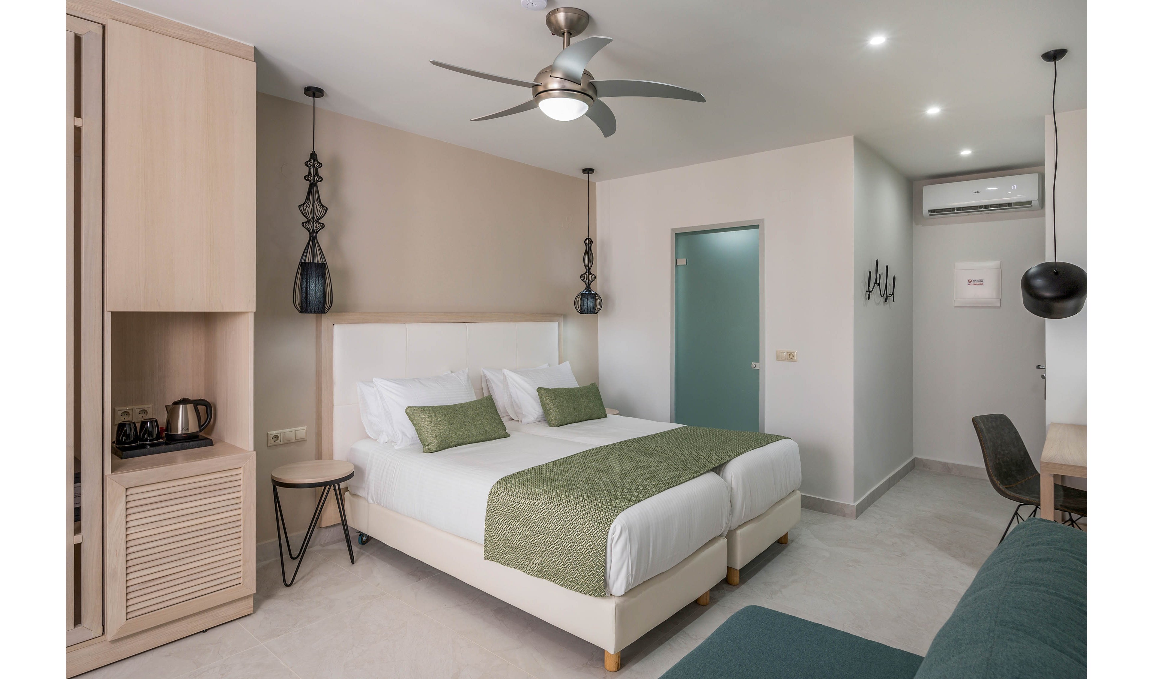 Contemporary hotel room featuring a double bed with white linens and green throws and cushions. The space includes built-in wooden storage, a small side table, and a desk with a chair. A ceiling fan and recessed lighting brighten the neutral-toned interior. A sofa in the foreground and a visible air conditioning unit enhance comfort in this clean, minimalist setting.