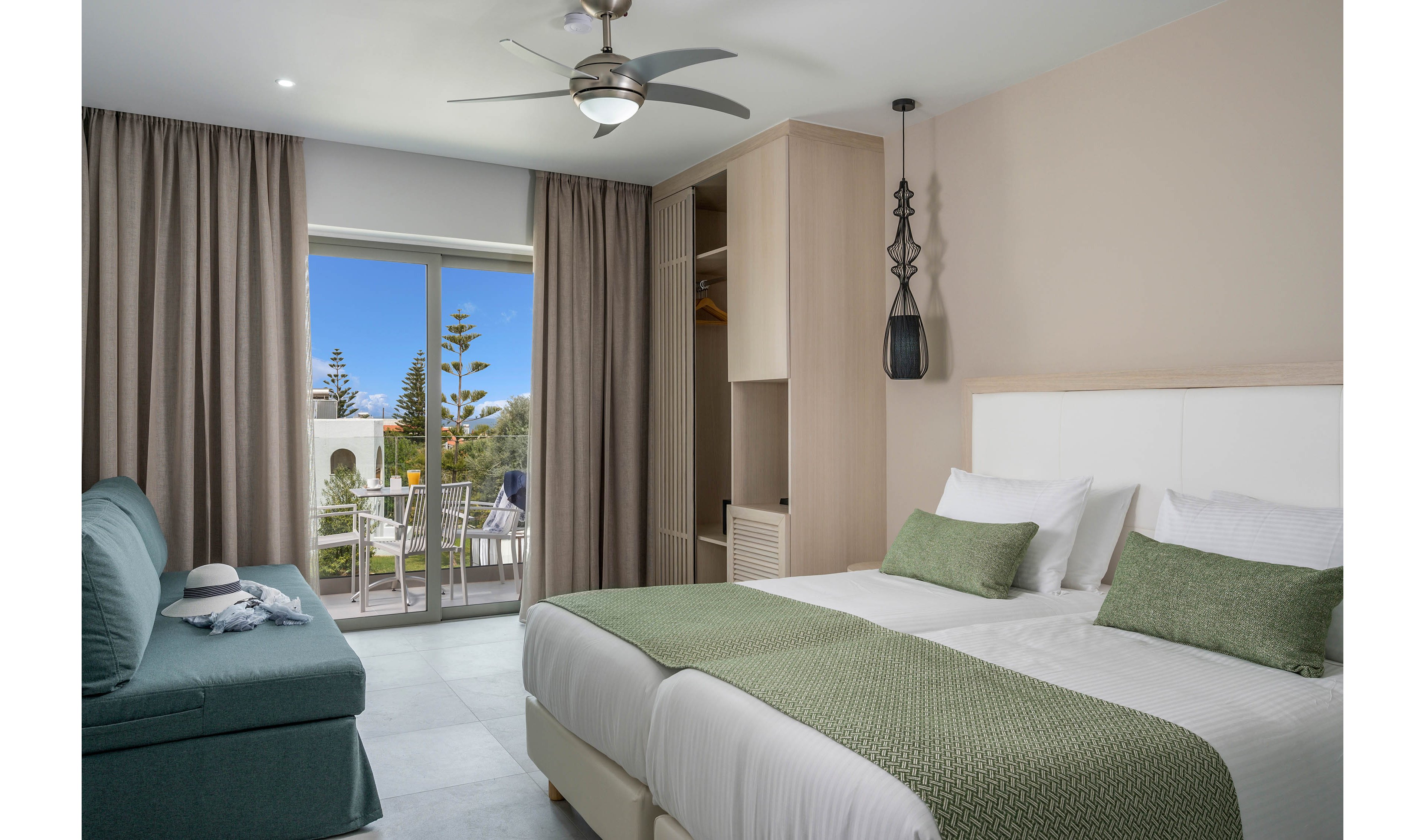 Contemporary hotel room featuring a large double bed with green accents and a matching sofa sits beside a sliding glass door. Neutral walls and floor tiles create a clean, minimalist look, complemented by long curtains and built-in wooden wardrobe. A ceiling fan and pendant light add modern touches to the interior. Outside, a balcony with chairs overlooks a garden area with trees under a bright blue sky.