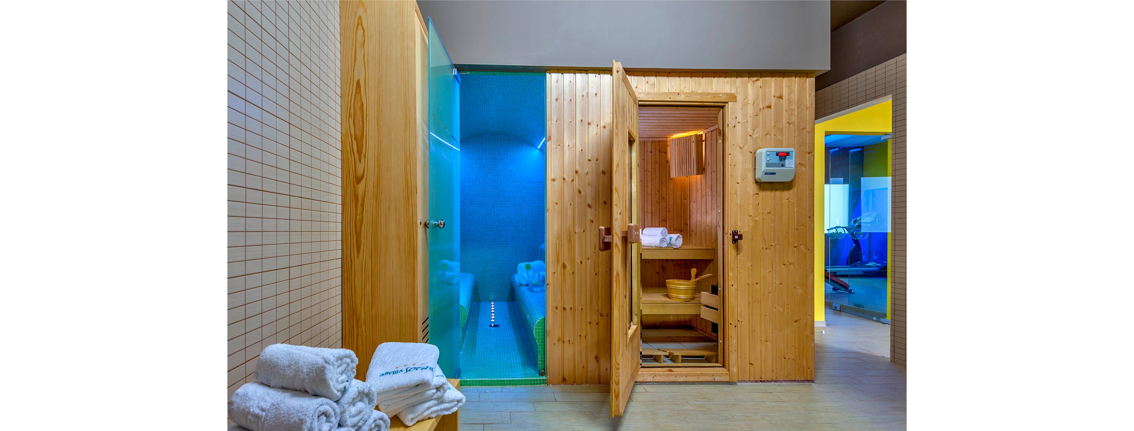 Interior view of a spa area featuring a wooden sauna cabin and an adjacent blue-lit steam room. Rolled white towels are neatly stacked on a bench in the foreground, emphasizing cleanliness and relaxation. The steam room glows with cool blue lighting, while the sauna emits a warm yellow light through its open door. Additional fitness or wellness spaces are visible through doorways in the background, suggesting a full-service wellness facility.