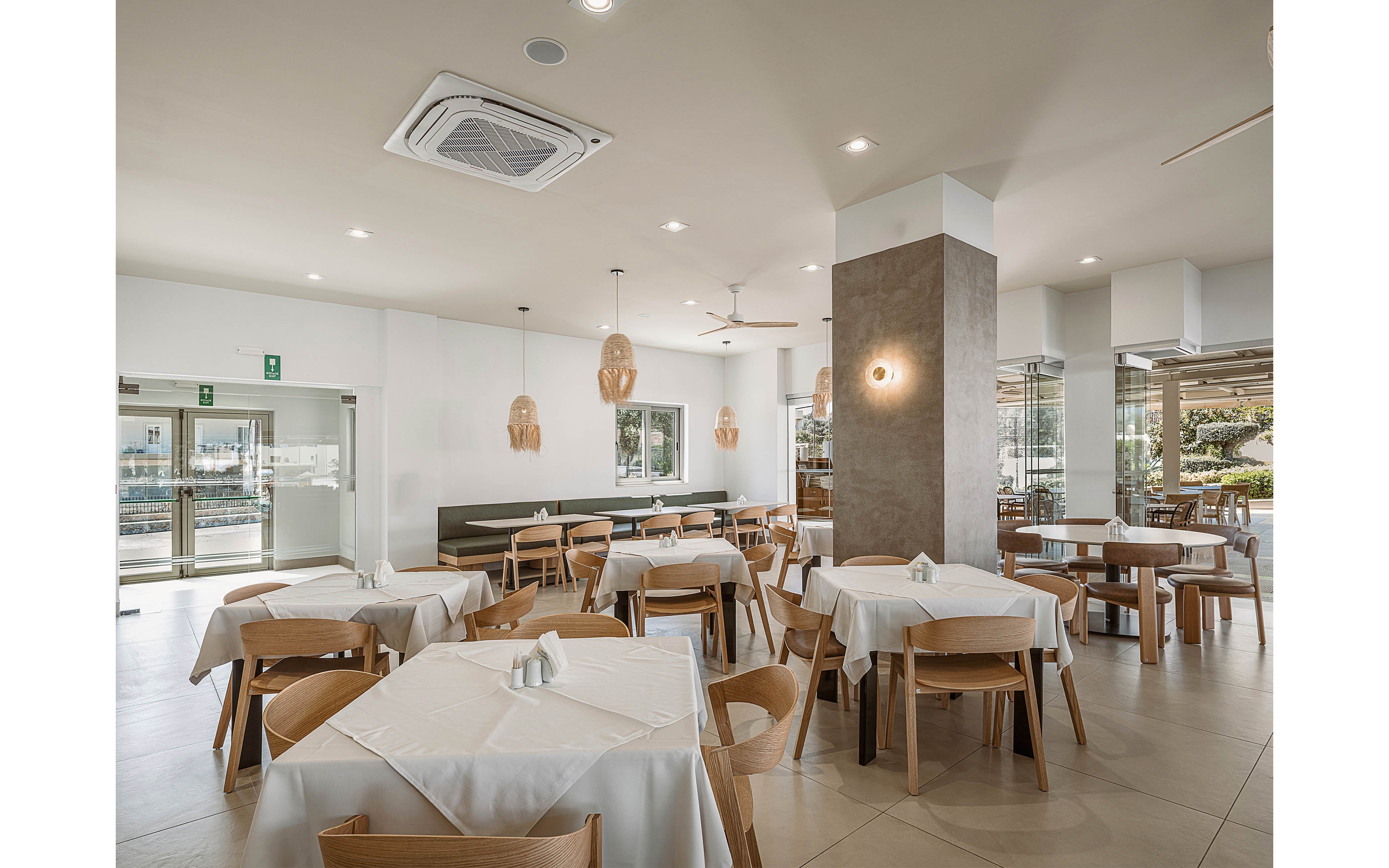 Spacious restaurant dining area featuring light wooden chairs and tables covered with white tablecloths. The room is bright and airy, with large windows and glass doors letting in natural light. Contemporary pendant lights and wall sconces add warm illumination to the minimalist decor. The overall atmosphere is clean, modern, and welcoming, suitable for casual or upscale dining.
