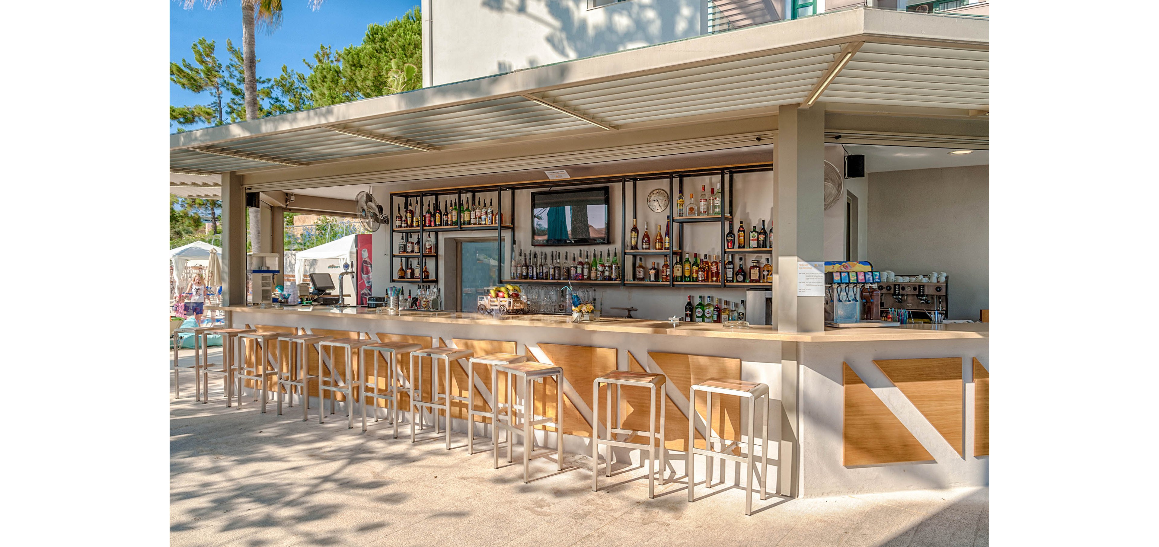 An open-air bar features a long counter lined with wooden stools and a wide selection of bottles on shelves behind the bartender. A man and woman sit at the bar, chatting and enjoying drinks in a relaxed resort setting. The bar structure is modern with clean lines, neutral colors, and a shaded roof. Sunlight, palm trees, and poolside lounge areas in the background create a bright, vacation atmosphere.